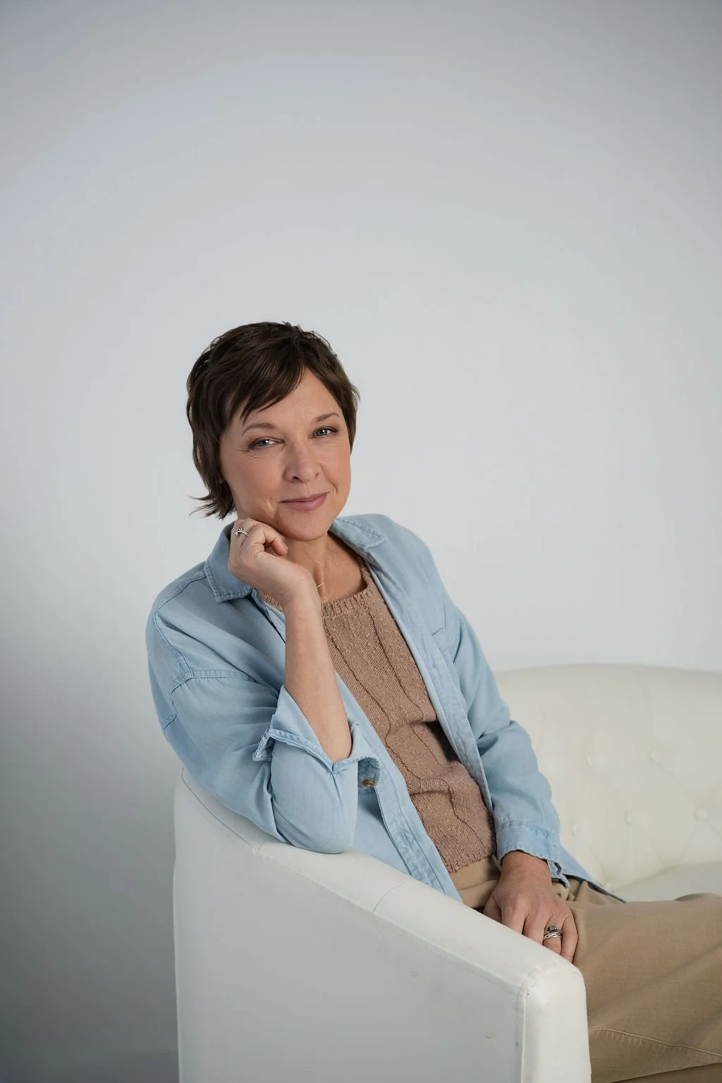 A woman with short brown hair, wearing a beige sweater and a light blue denim jacket, sitting on a white chair against a plain white background, with her hand resting on her chin and looking at the camera.
