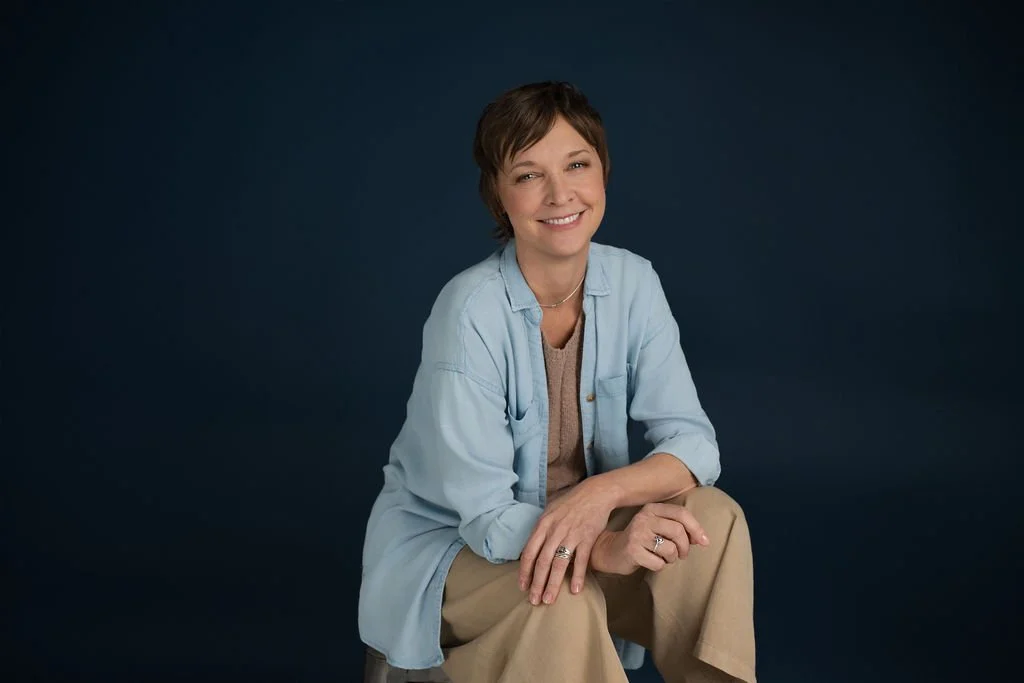 A woman with short brown hair smiling and sitting against a dark background, wearing a light blue shirt over a beige top and khaki pants.