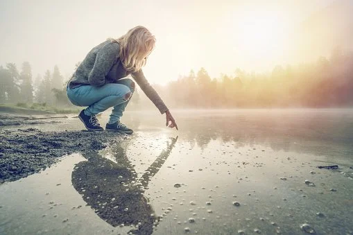Person crouching by a calm body of water during sunrise or sunset, touching the water with their hand.