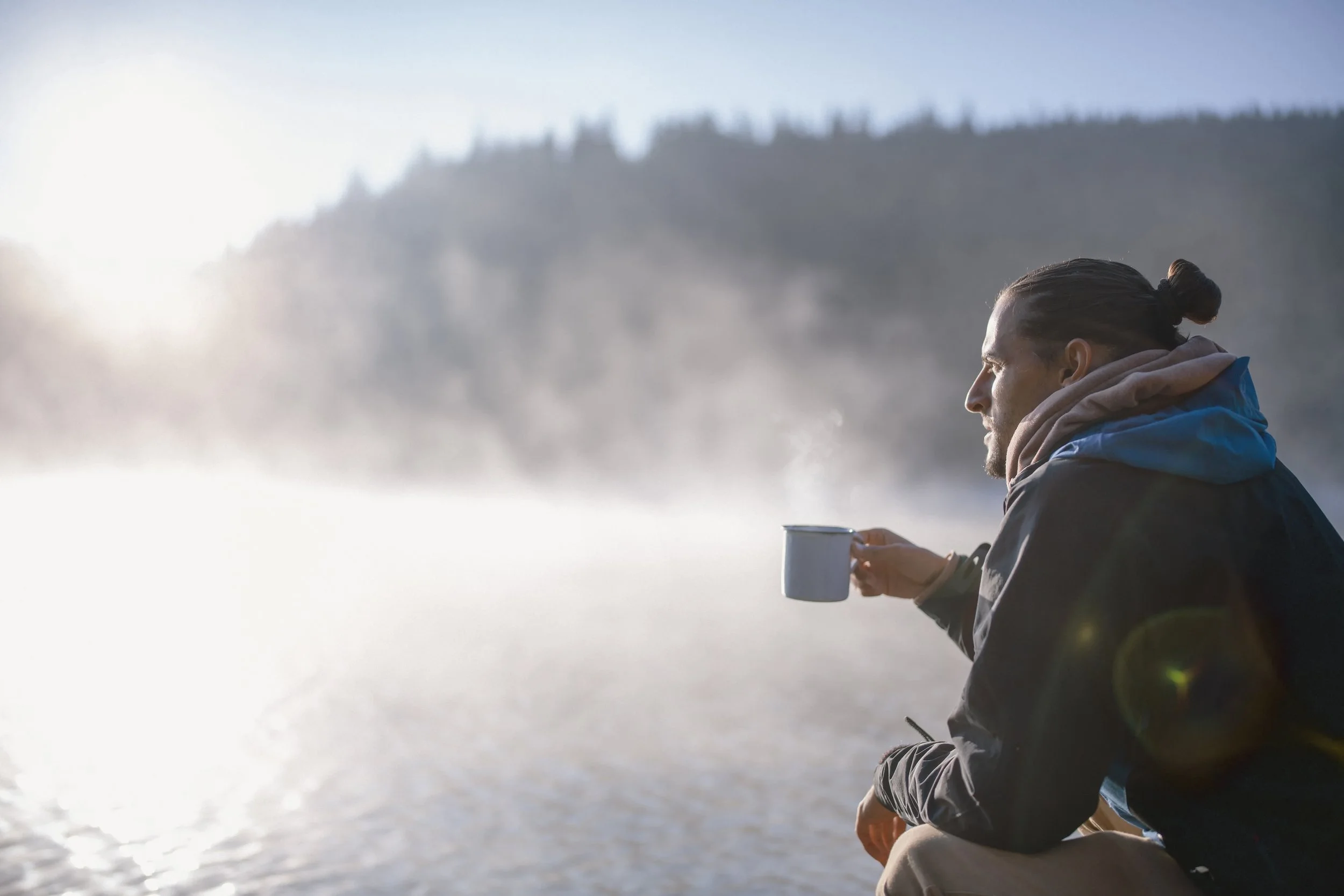 Man sitting outdoors near water, holding a mug, wearing a jacket and hoodie, with mountains and sunlight in the background.