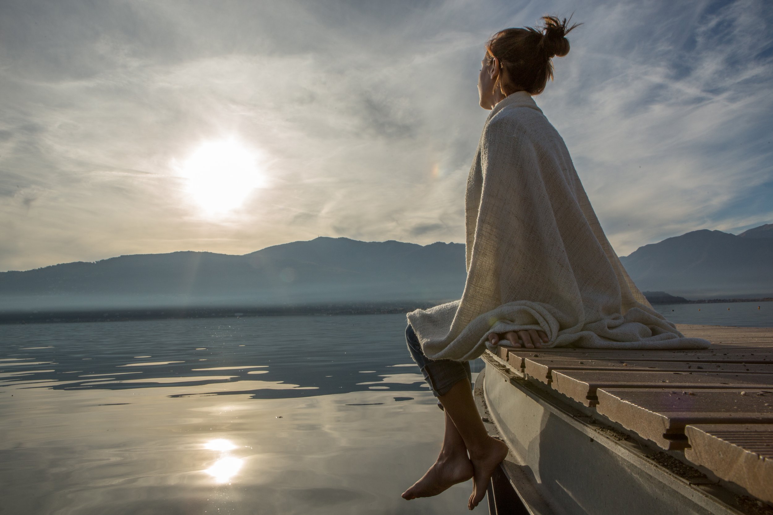 A woman sitting on the edge of a boat on a lake, wrapped in a blanket, gazing at the sun and mountains in the distance, during sunrise or sunset.