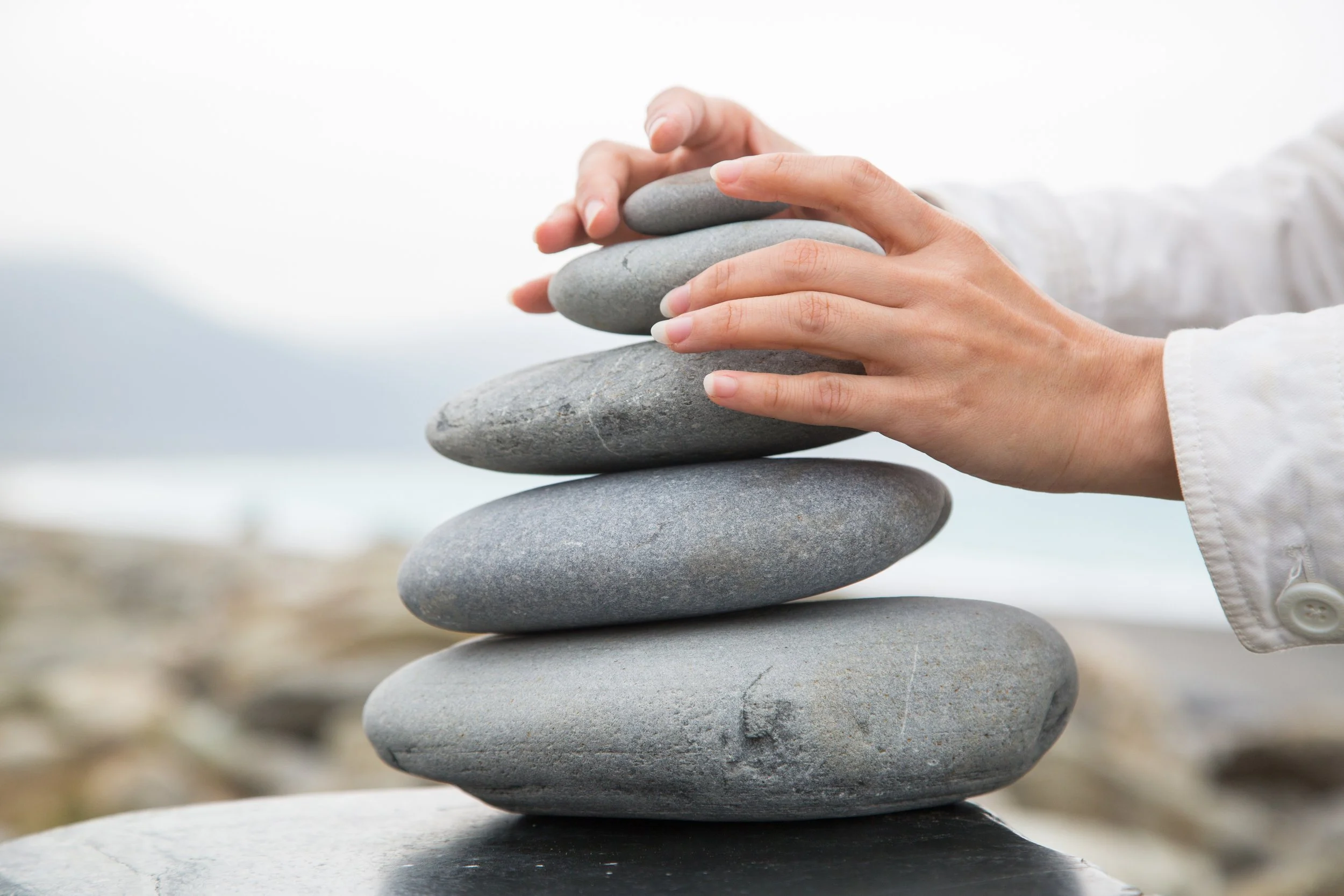 Person stacking smooth gray stones on a black surface outdoors, with a blurred beach and ocean in the background.