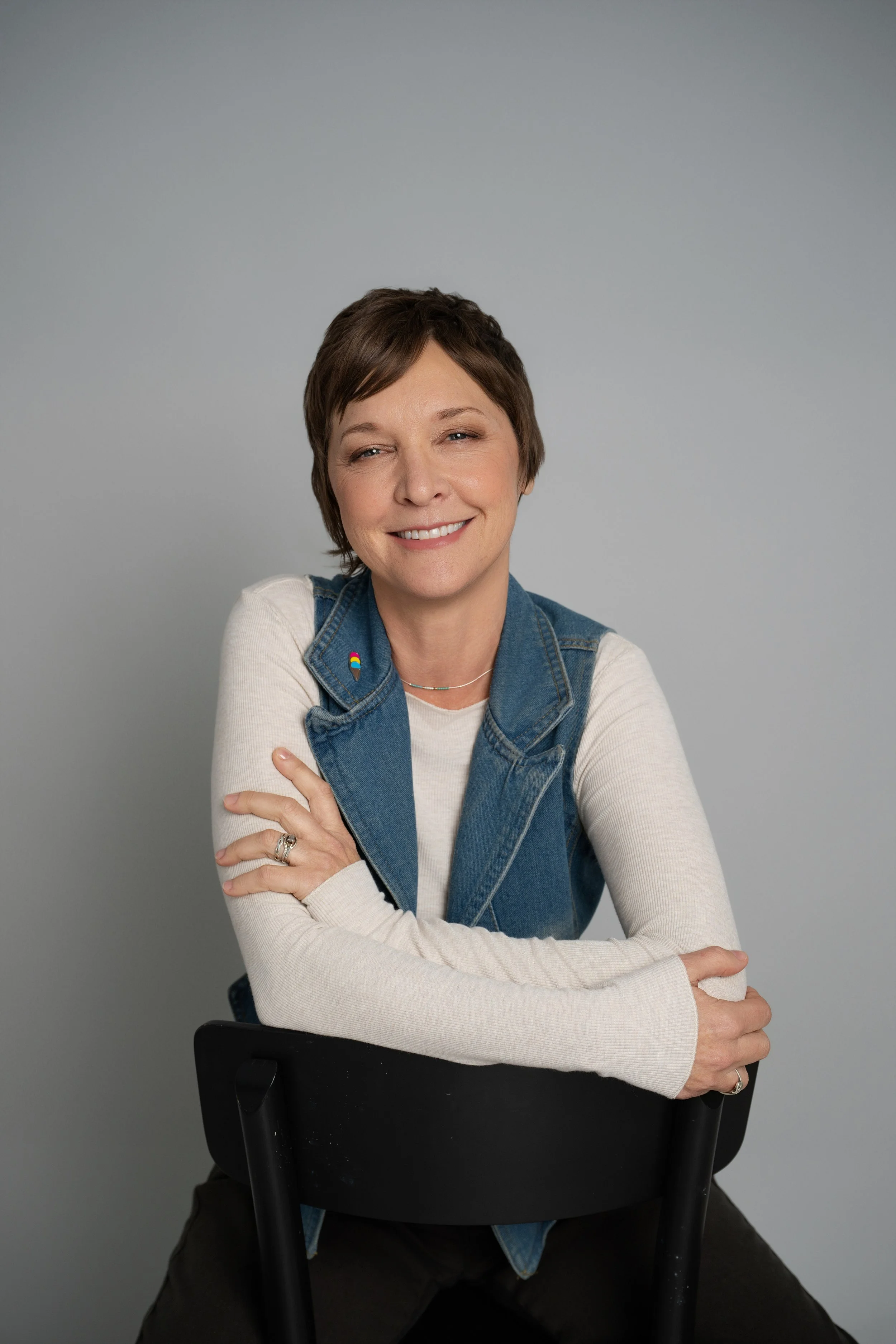 Smiling woman with short brown hair sitting on a black chair against a plain gray background, wearing a light-colored long sleeve shirt and a sleeveless denim vest with a rainbow-colored pin.