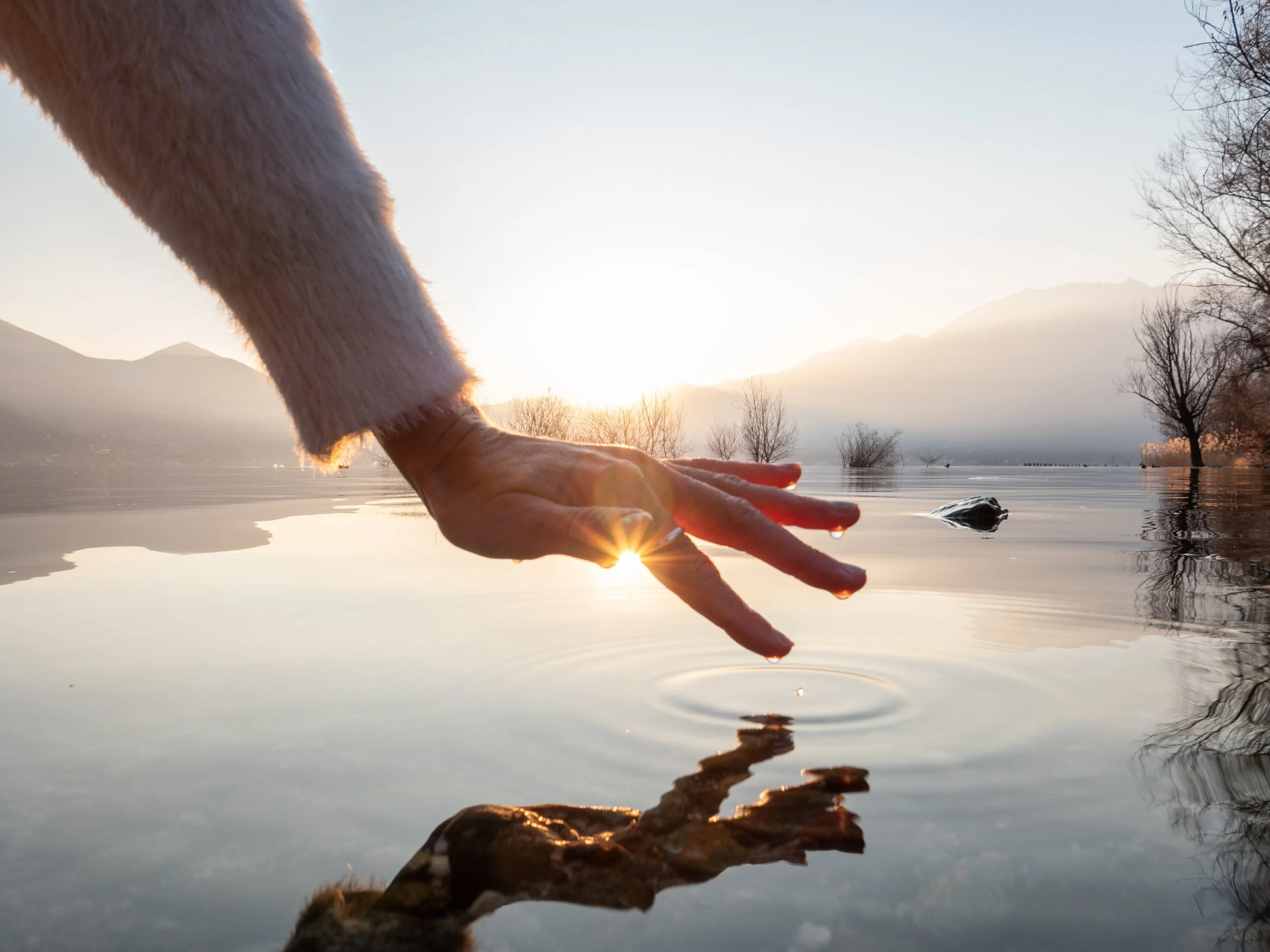 Person's hand with fingers extended, touching the surface of a calm lake during sunrise or sunset, with mountains and leafless trees in the background.
