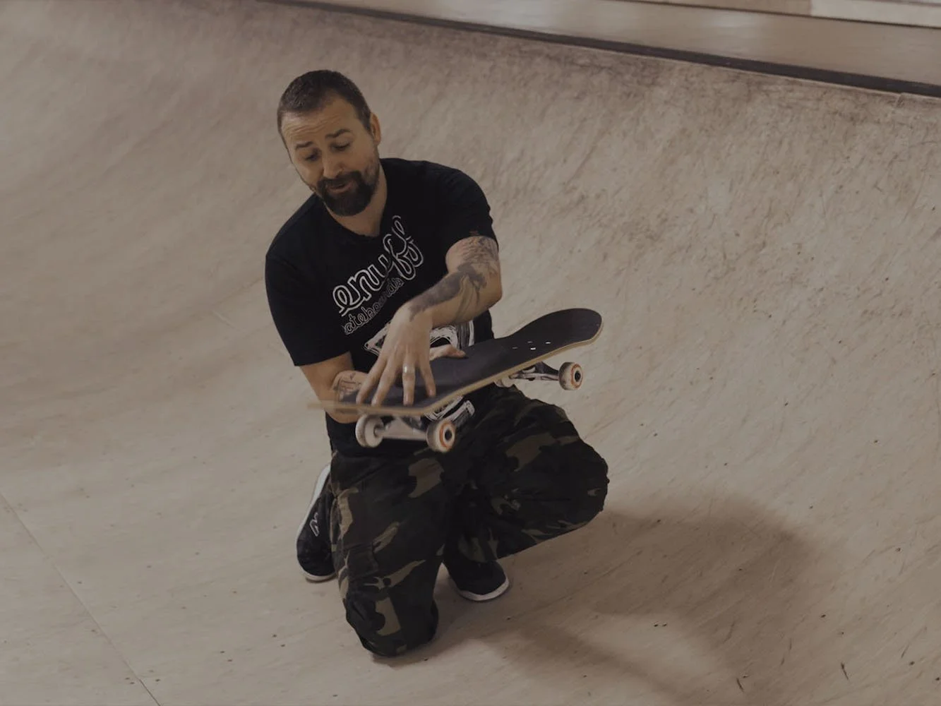 A man with a beard and tattoos kneeling on one knee in a skate park, holding a skateboard.