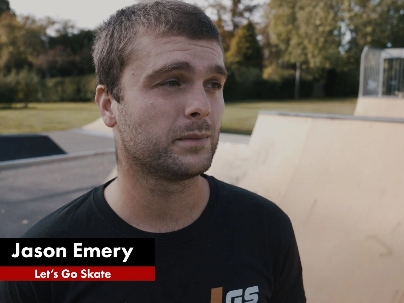 A man with short brown hair and facial hair, wearing a black shirt, standing at a skate park with ramps and trees in the background.