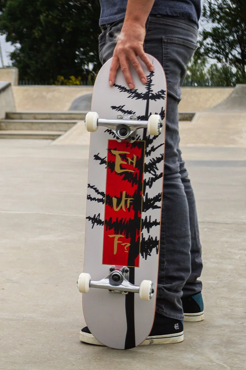 Person standing at a skatepark holding a skateboard with a tree and lightning bolt graphic design.
