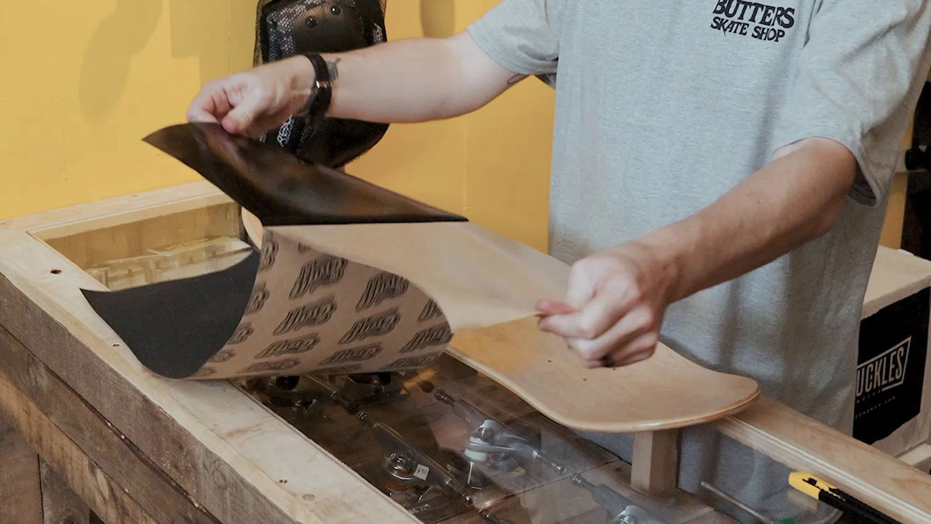 A person is peeling off protective paper from a skateboard deck in a workshop with wood and skateboarding tools visible.