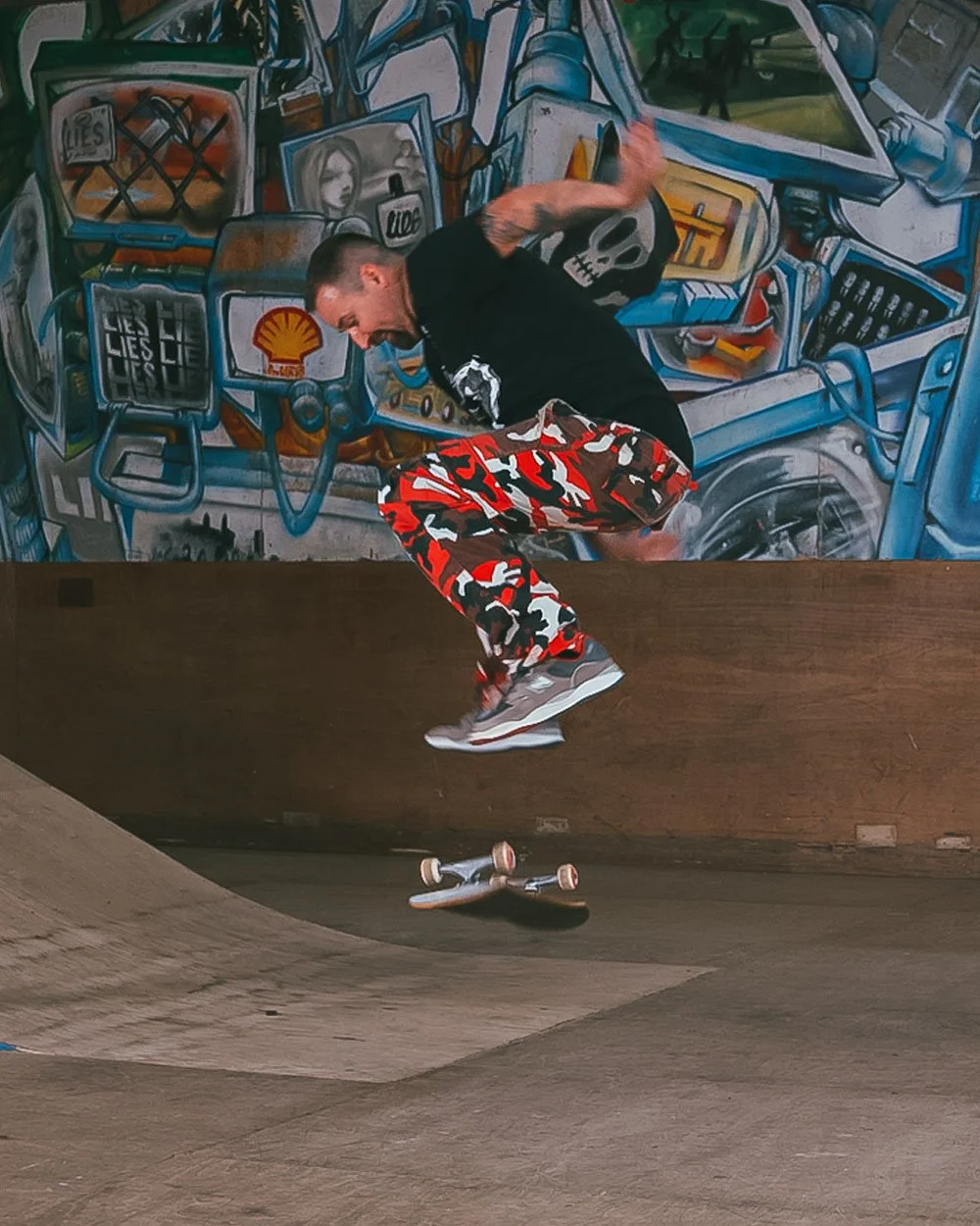 Skateboarder performing a trick in an indoor skate park with a graffiti mural on the wall behind him.