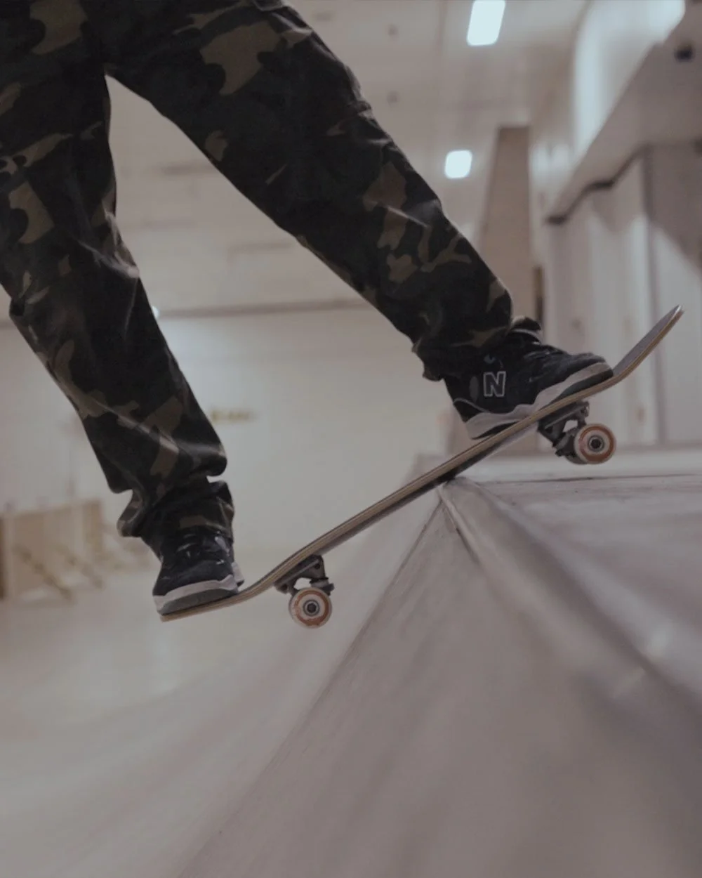 Skateboarder wearing camouflage pants and black sneakers performing a trick on a ramp in an indoor skatepark.