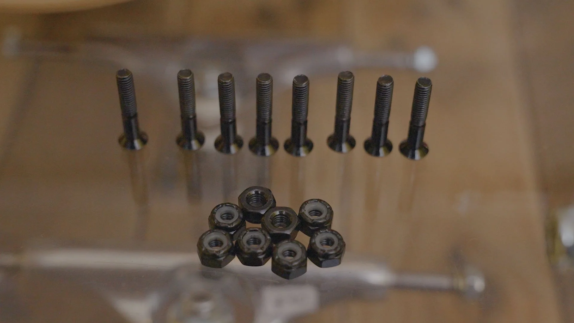 A metallic surface with ten black bolts standing upright in a row and eight black nuts placed in a cluster in the foreground.