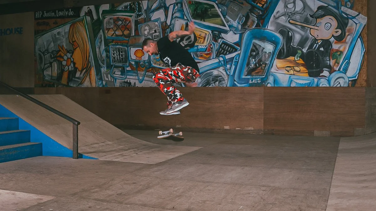 Skateboarder performing a trick in an indoor skate park with colorful graffiti art on the wall behind him.