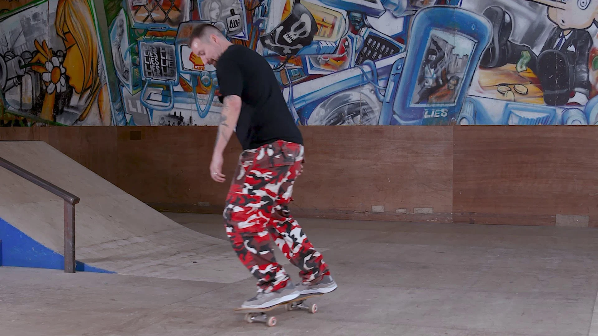 A man skateboarding at an indoor skate park with a colorful graffiti mural on the wall behind him.