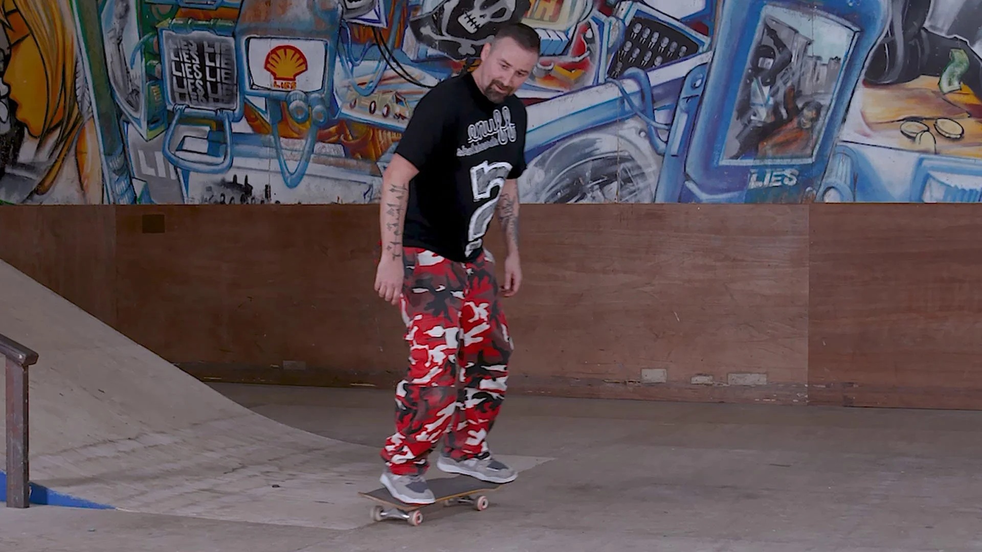 A man skateboarding inside a skate park with murals on the wall behind him, wearing a black t-shirt with a white logo and red camouflage pants.