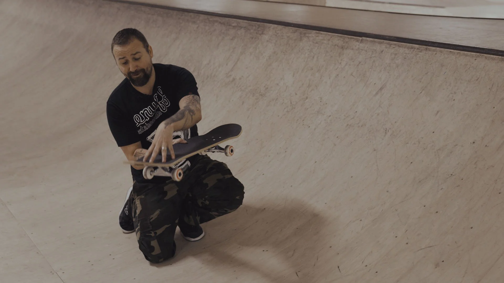 A man with tattoos on his arms and beard, wearing a black t-shirt and camouflage pants, is kneeling while holding a skateboard inside a skatepark.