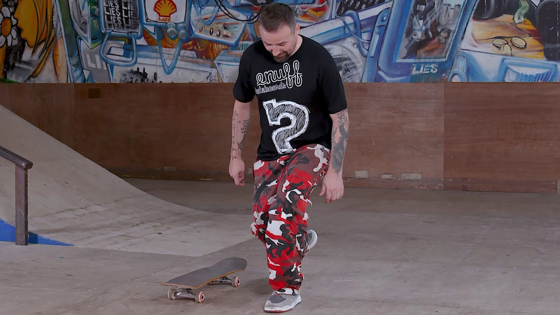 A man with tattoos dressed in a black T-shirt with a large number 2 and camo-style red, black, and white pants, skateboarding at an indoor skatepark.