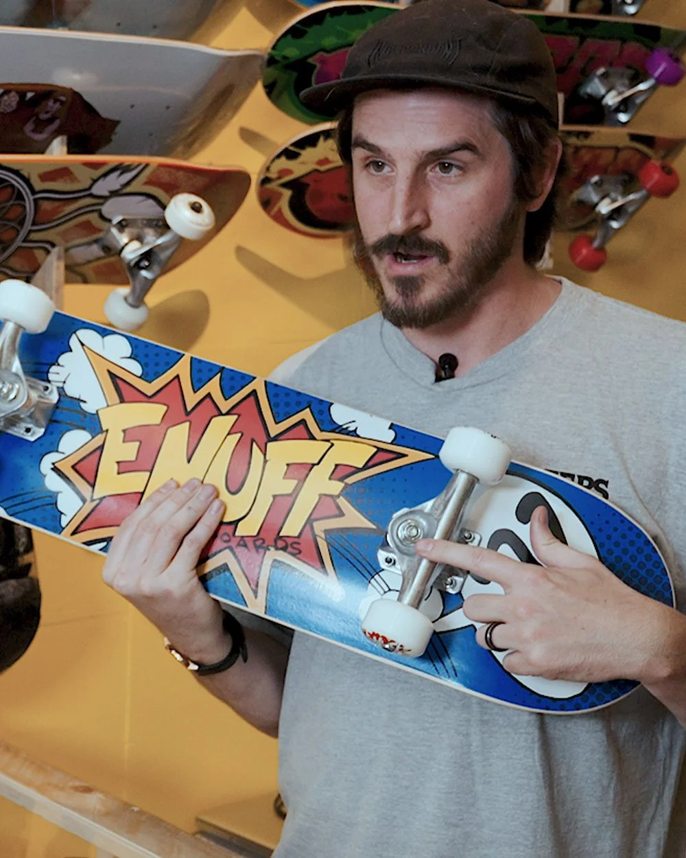 A man with a beard and mustache wearing a black cap and gray shirt holding a skateboard with a comic book style design that says 'ENUFF' in large letters, inside a store with skateboards displayed on the wall behind him.