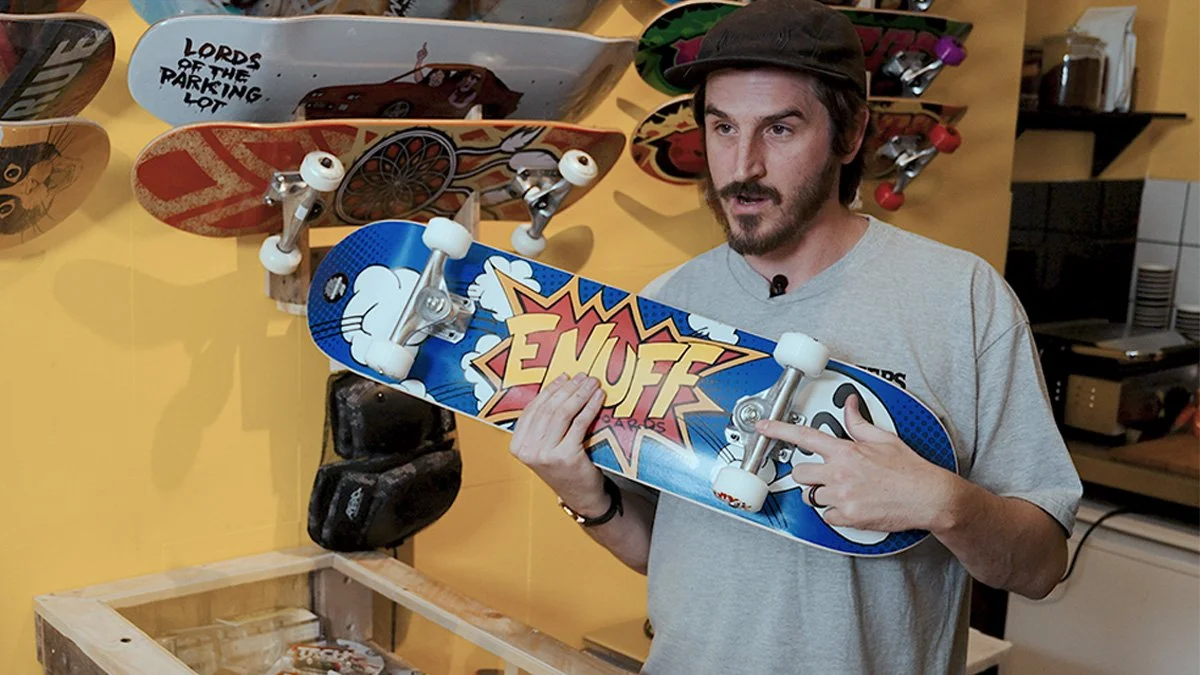Man holding a skateboard with a comic-style 'Emuff' graphic, in a skate shop with other skateboards on display.