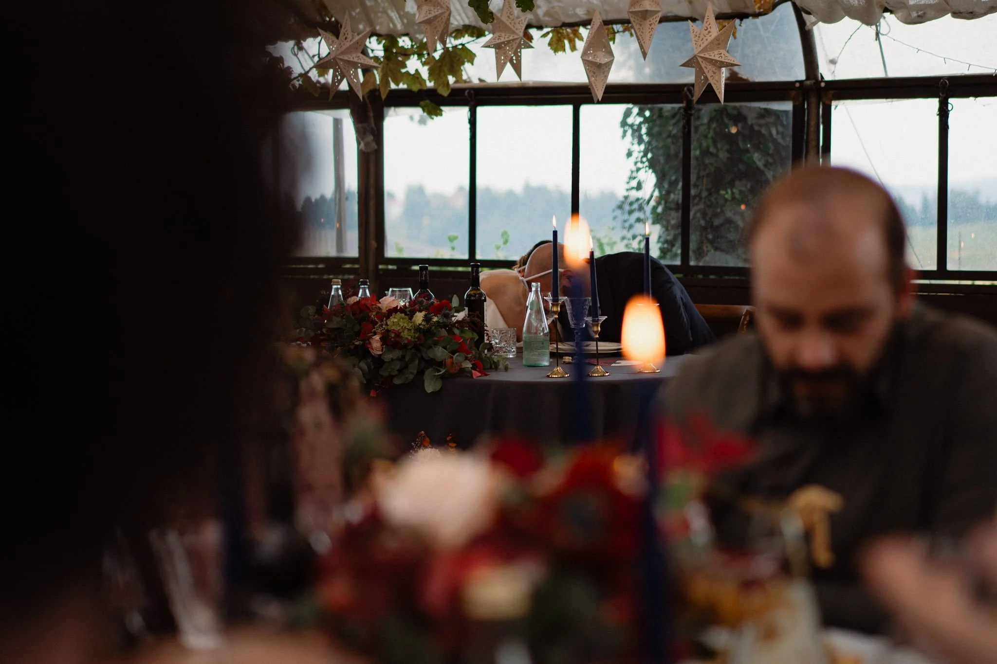Una scena di un pranzo di festa in un ambiente interno con luci soffuse, decorazioni di stelle sospese al soffitto, tavolo apparecchiato con fiori, candeline e bottiglie di vino. Due persone sono sedute al tavolo, una con la testa reclinata e un'altr