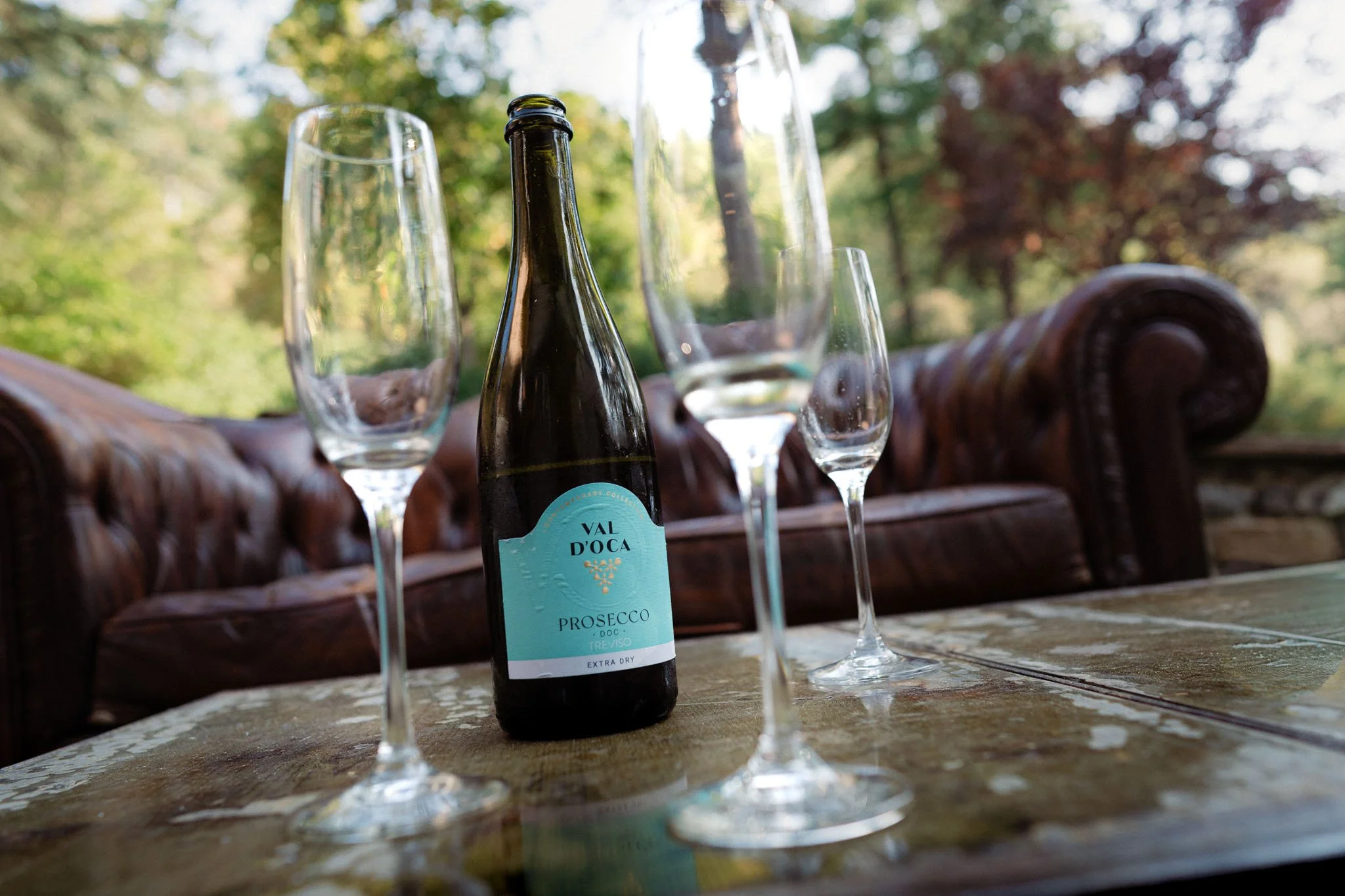 Bottiglia di Prosecco Val D'Oca e quattro flute da champagne vuote su un tavolo rustico all'aperto con sfondo di alberi e natura.