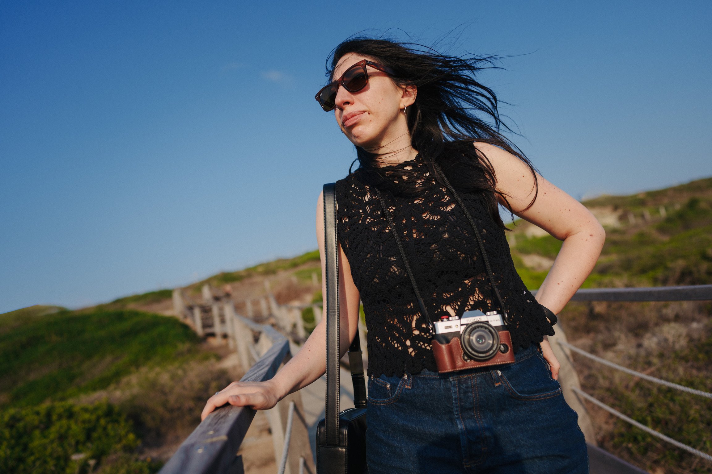 Una donna con occhiali da sole e camicia all'aperto sulla scogliera, con una macchina fotografica appesa al collo, che si appoggia sul parapetto di una passerella in legno sotto un cielo blu.