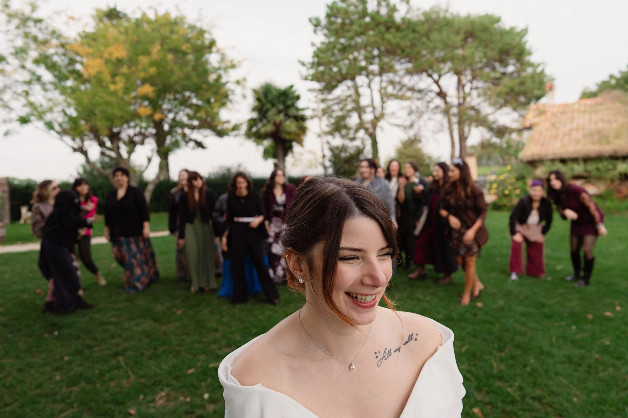 Una giovane donna sorridente con capelli castani e tatuaggio sulla spalla, in un abito bianco, in primo piano. Sullo sfondo molte persone, principalmente donne, che ridono e si divertono in un giardino con alberi e casette, durante un evento all'aper