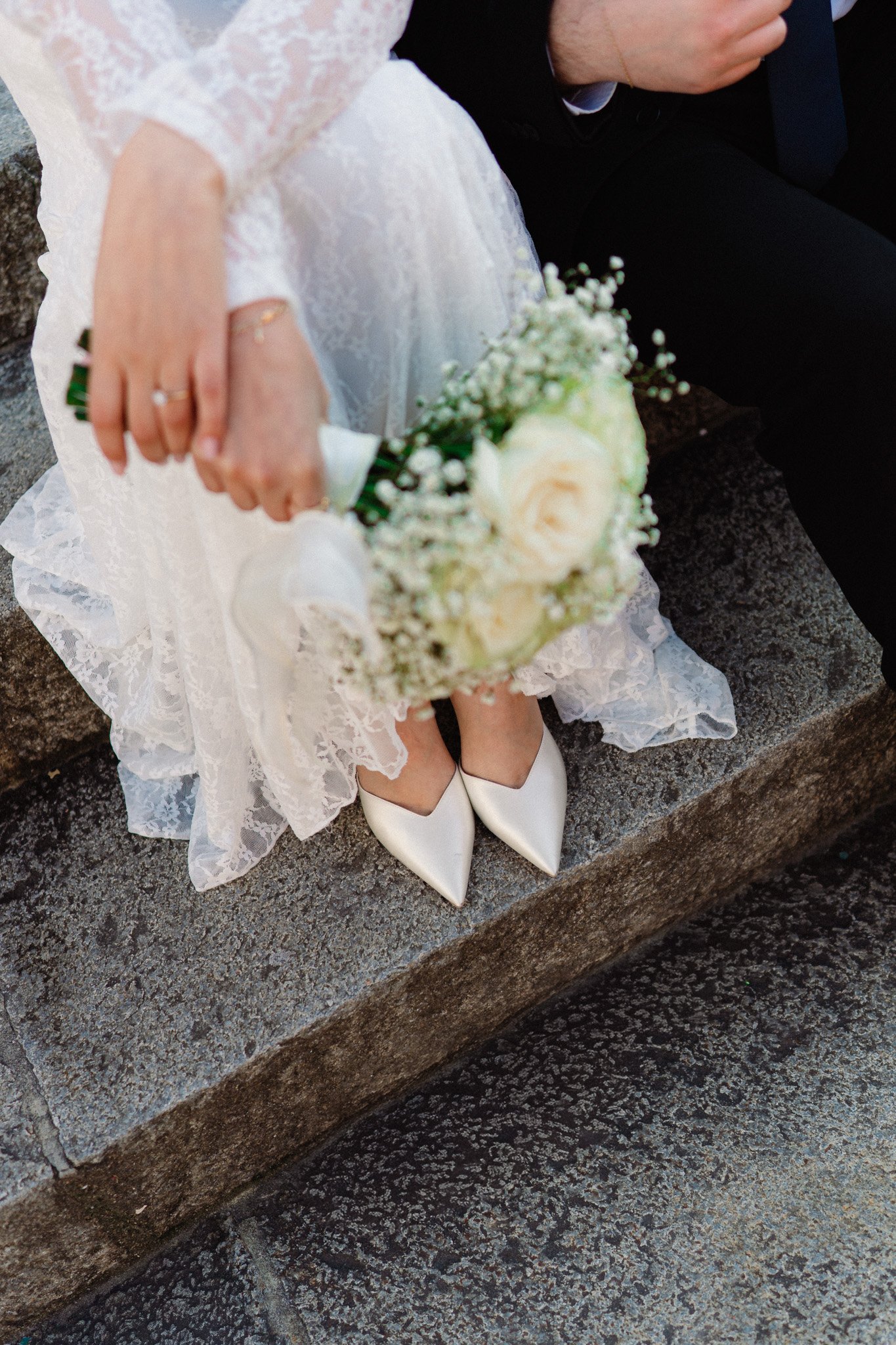 Dettaglio di una sposa in abito bianco con scarpe a punta bianca, seduta su un muretto di pietra, che tiene un bouquet di fiori bianchi, accanto a una persona in abito scuro.