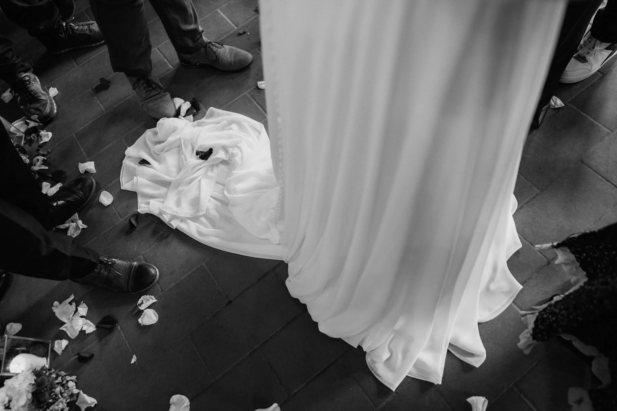 Dettaglio di un'abito da sposa disteso sul pavimento, circondato da pedali di fiori e persone in piedi intorno, in una scena in bianco e nero.