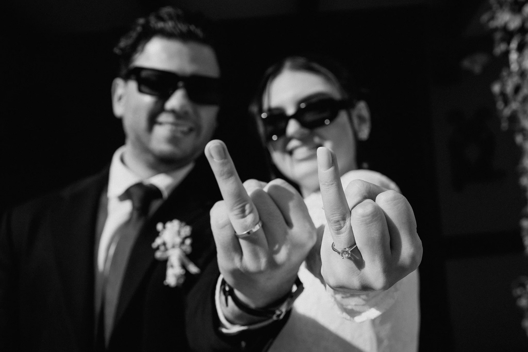 Una coppia di persone indossa occhiali da sole e mostra le mani con le dita con la fede nuziale, sorridendo.