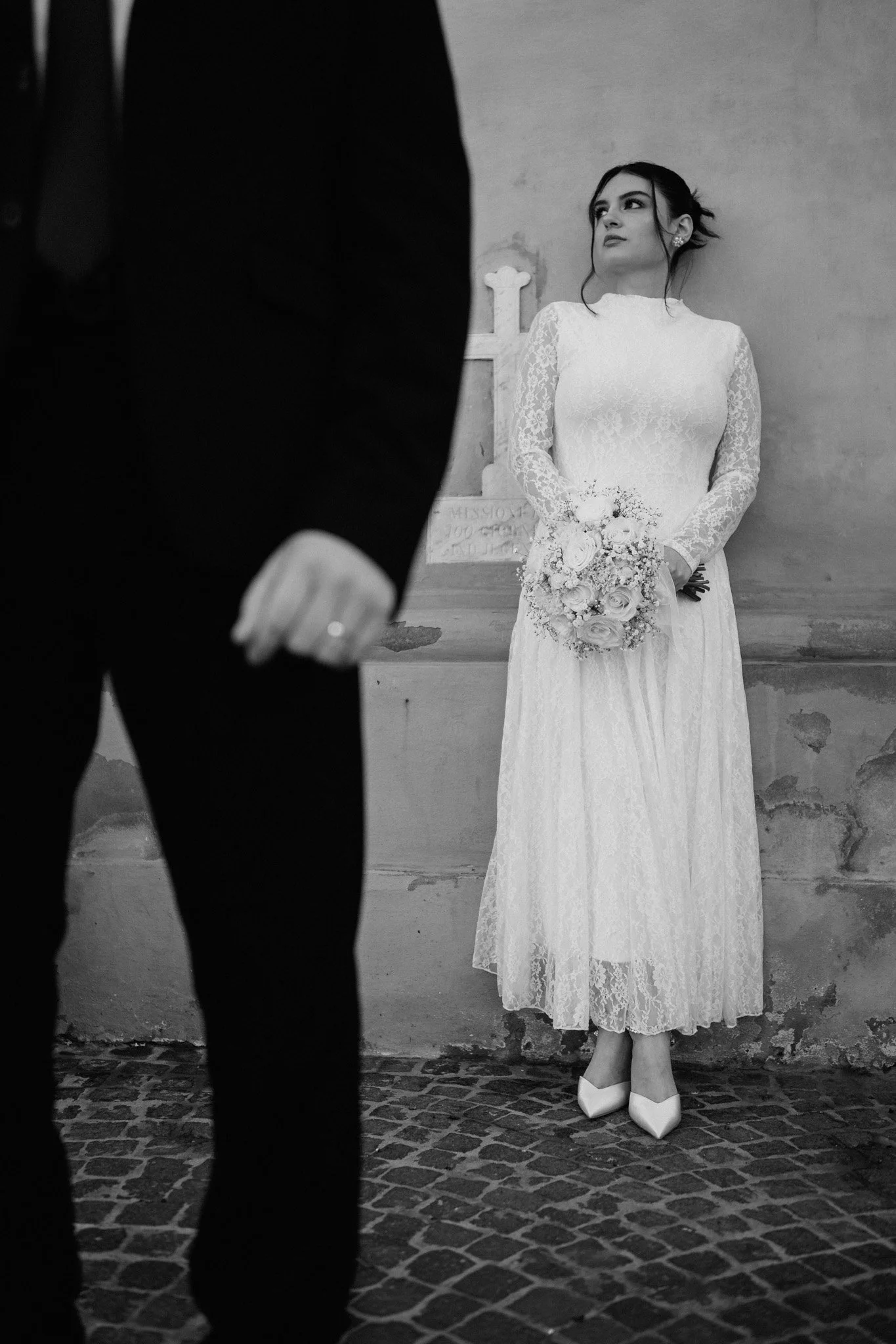 Una sposa in abito bianco con velo, durante un matrimoni vicino Bologna che tiene un bouquet di fiori, si trova contro un muro di pietra. Davanti a lei c'è una persona in abito nero, visibile solo parzialmente.