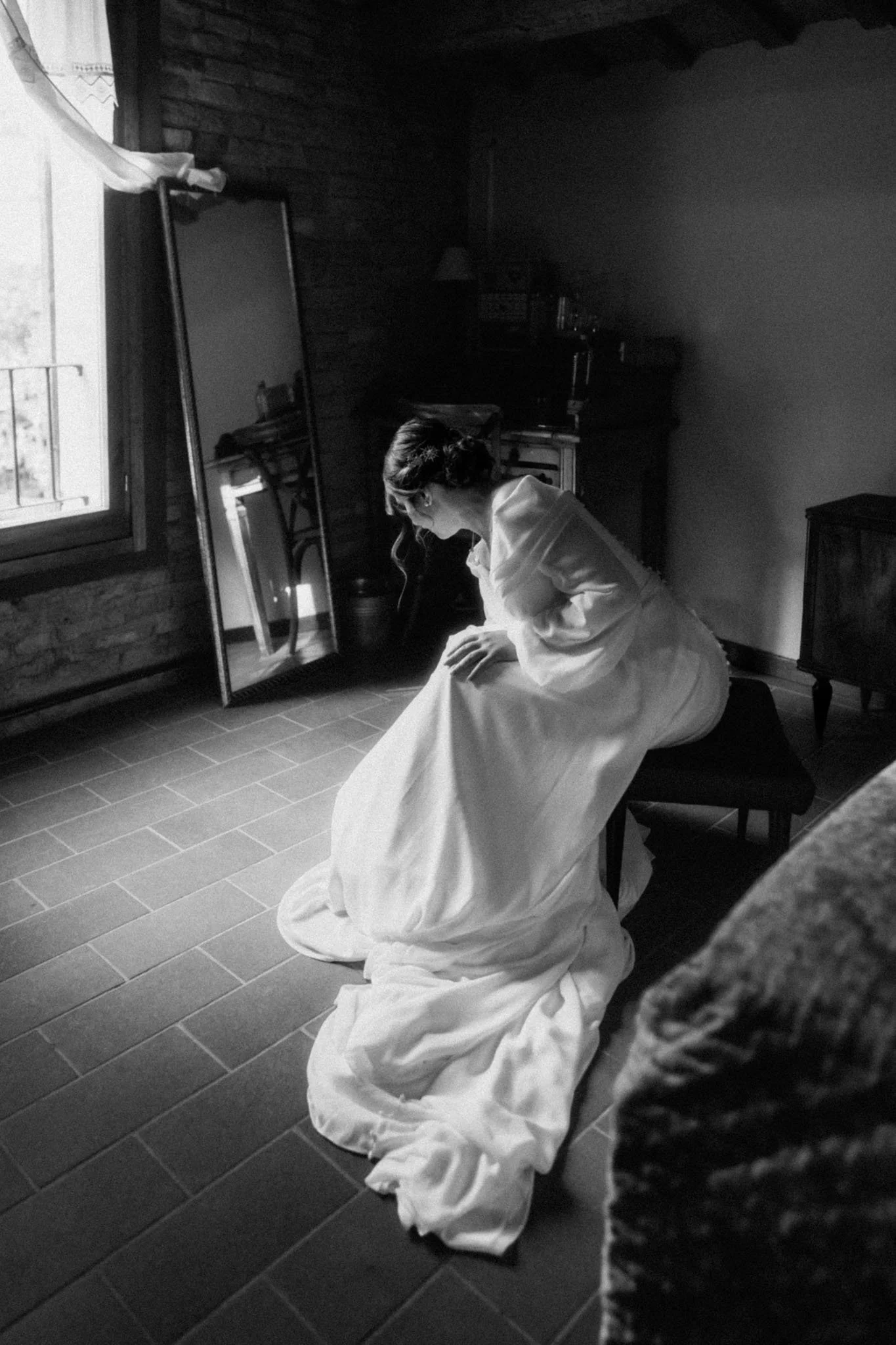 Donna seduta su uno sgabello, indossa un lungo vestito bianco e guarda un grande specchio in una stanza con pareti di legno e pavimento in piastrelle, illuminata dalla luce che entra da una finestra.