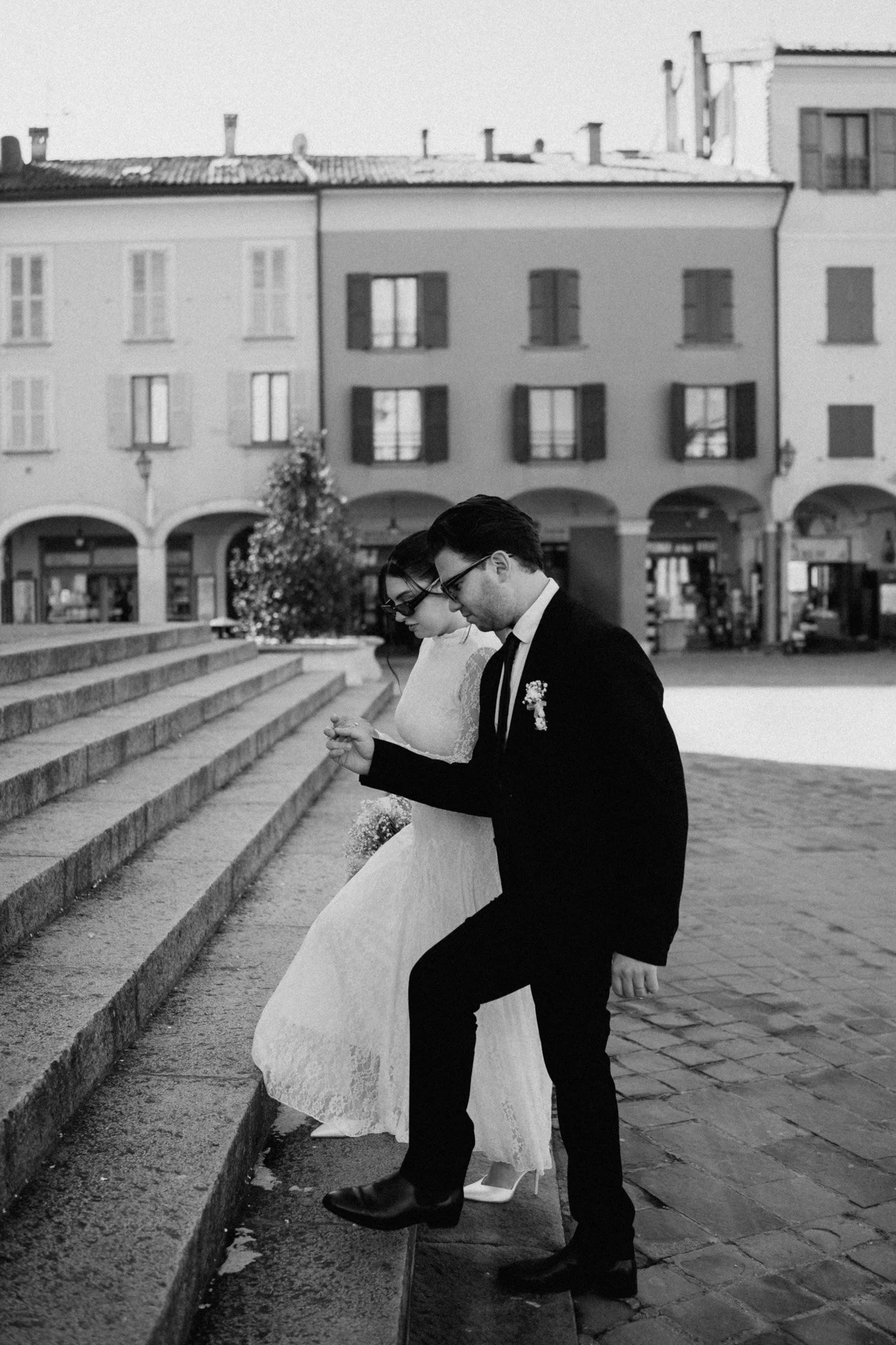 Una coppia di sposi si sta attraversando le scale all'aperto in un'area urbana, la donna indossa un abito da sposa bianco e alta, l'uomo indossa un abito nero con papillon, entrambi indossano occhiali da sole e sono in stile vintage in bianco e nero.