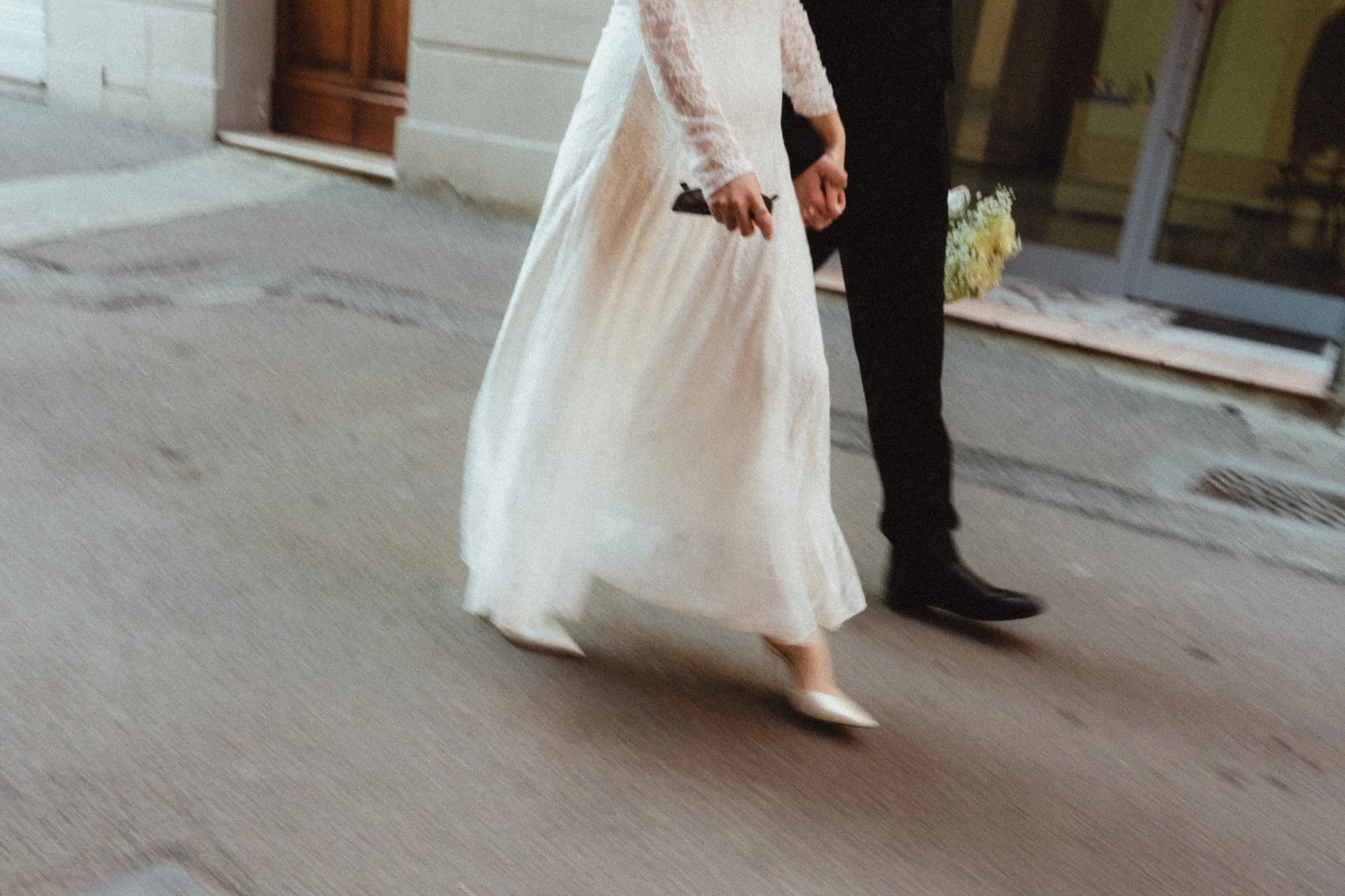 Due sposi a un matrimonio, una donna con abito bianco lungo e scarpe di tacco, e un uomo in abbigliamento scuro, camminano mano nella mano su un marciapiede.