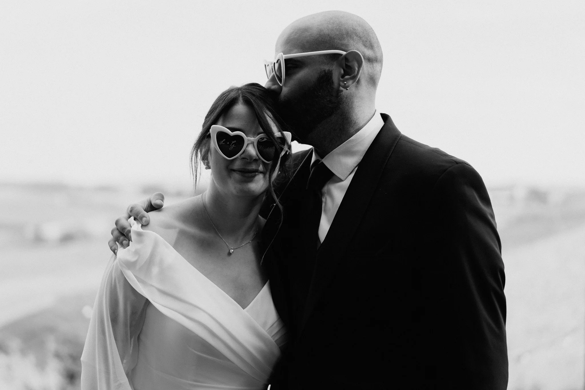 Una coppia si abbraccia, lei indossa occhiali a cuore e una gonna a drappeggio, lui in completo nero con camicia bianca.