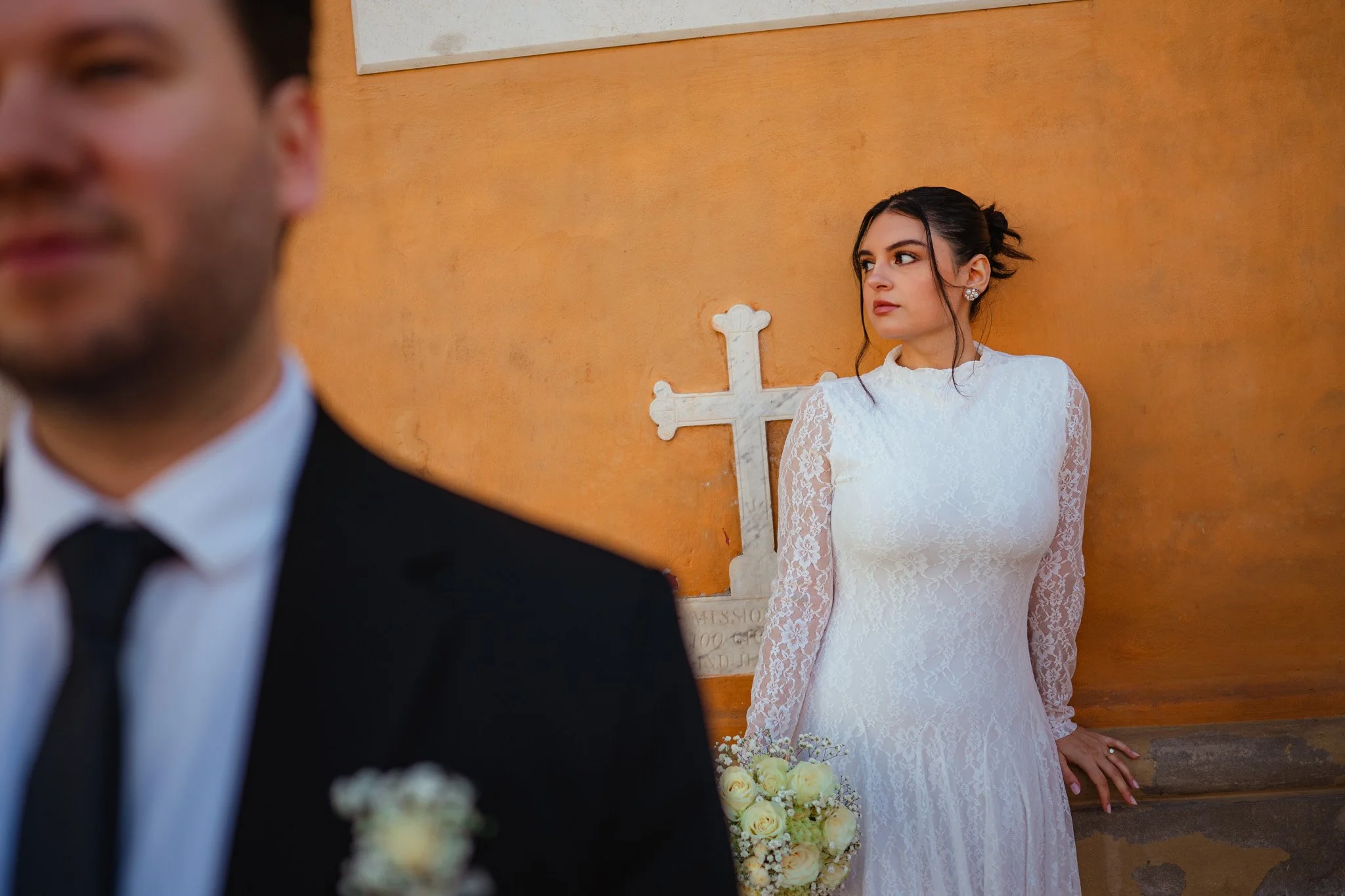 Una donna in abito da sposa bianco con ricami, che tiene un bouquet di fiori chiari, in piedi contro una parete di colore arancione con una croce in pietra alle sue spalle, in un contesto di matrimonio religioso.