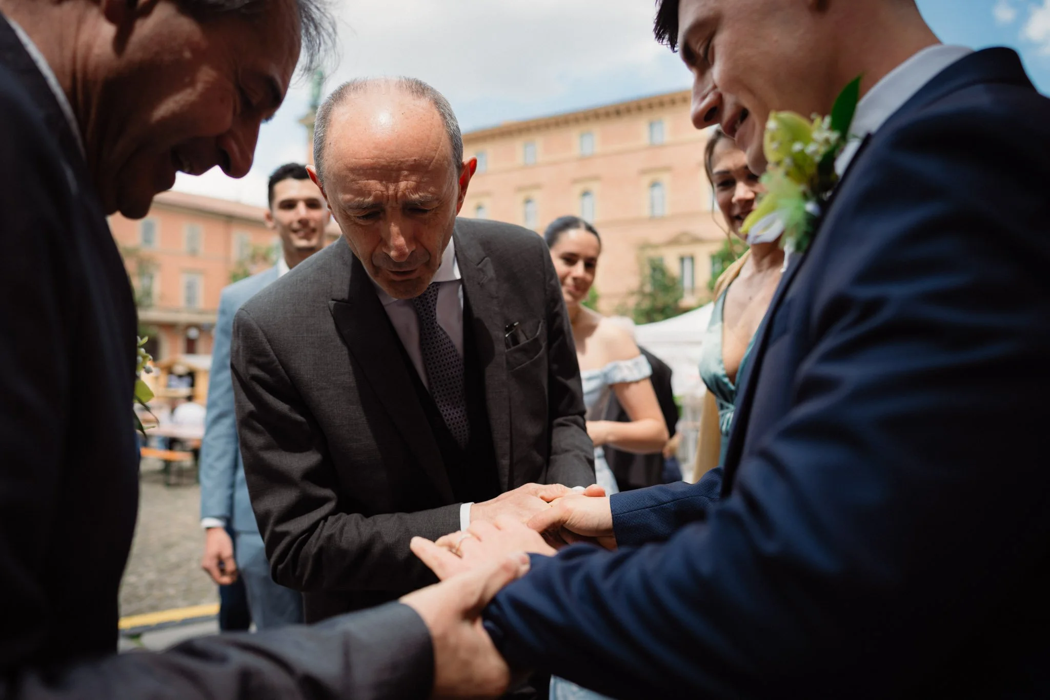 Una coppia di sposi si tiene per mano durante un matrimonio all'aperto, circondata da amici e parenti, in un cortile con edifici storici sullo sfondo.