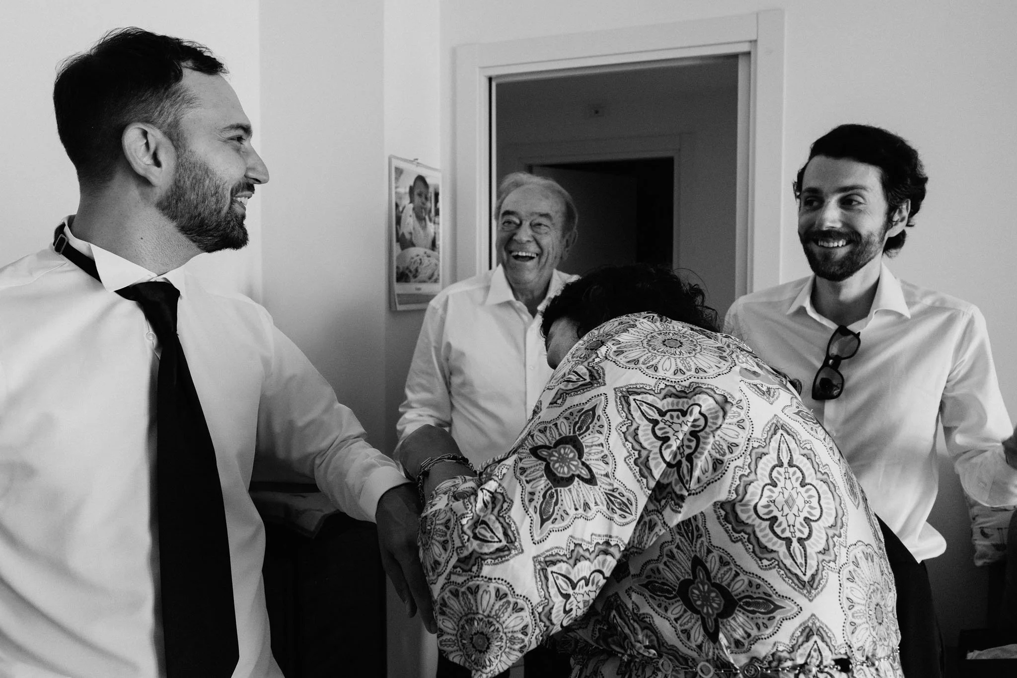 Gruppo di quattro persone che ridono e si scambiano un saluto affettuoso in una stanza, con una donna che si inchina verso un uomo e gli dà un bacio sulla mano, tutti sorridono.