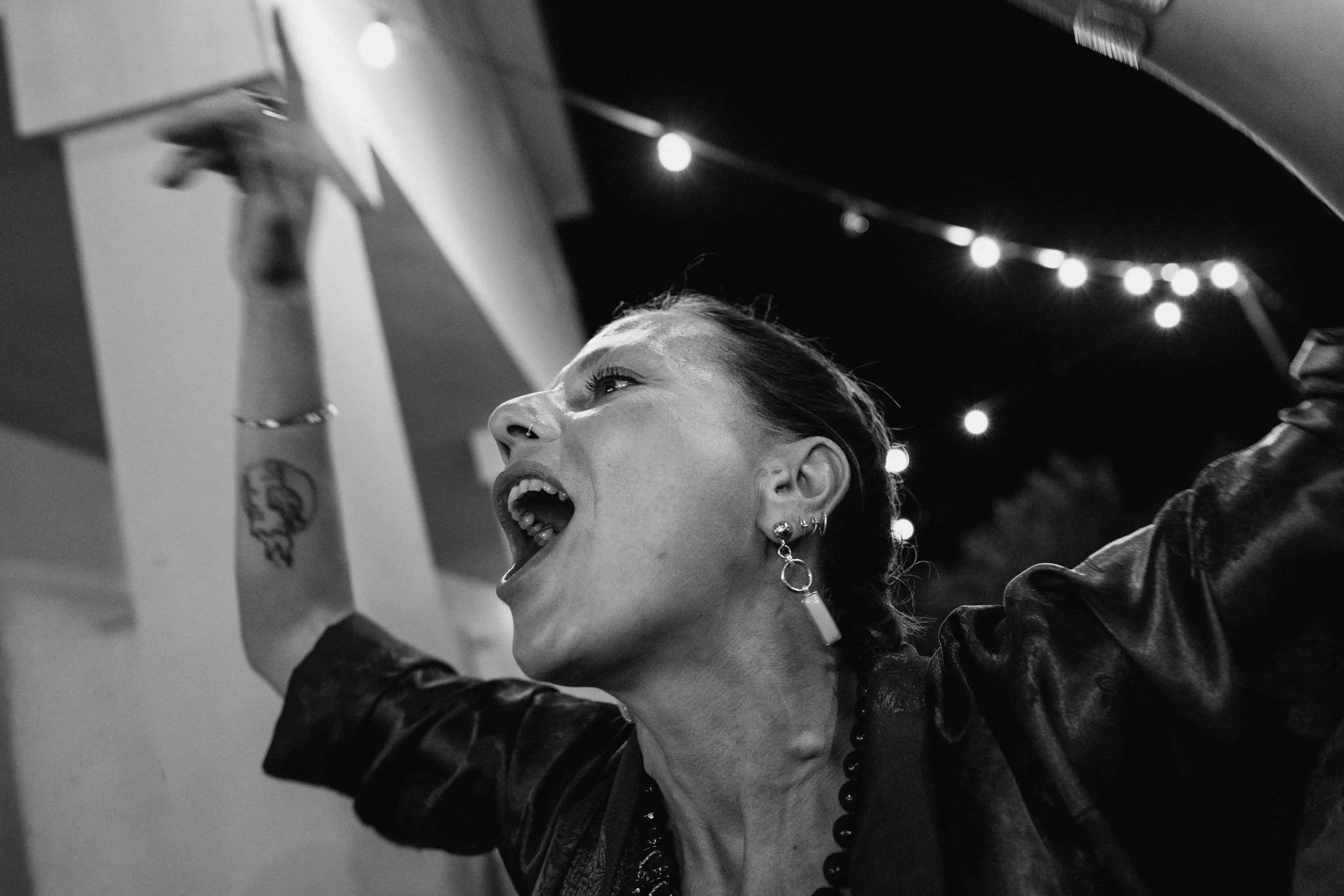 Una donna con capelli raccolti, tatuaggi e orecchini, sorride e alza le braccia in un ambiente con luci di festa.