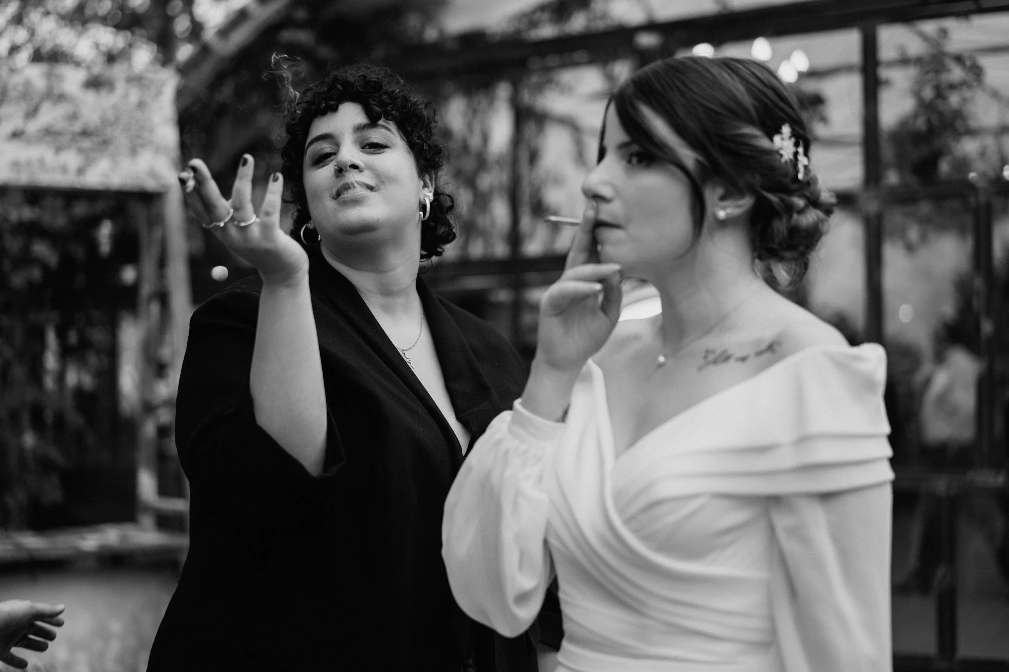 Due donne, una con capelli ricci e unghie nere, l'altra con capelli raccolti e tatuaggio sulla spalla, mentre fumano sigarette e parlano in un ambiente interno.