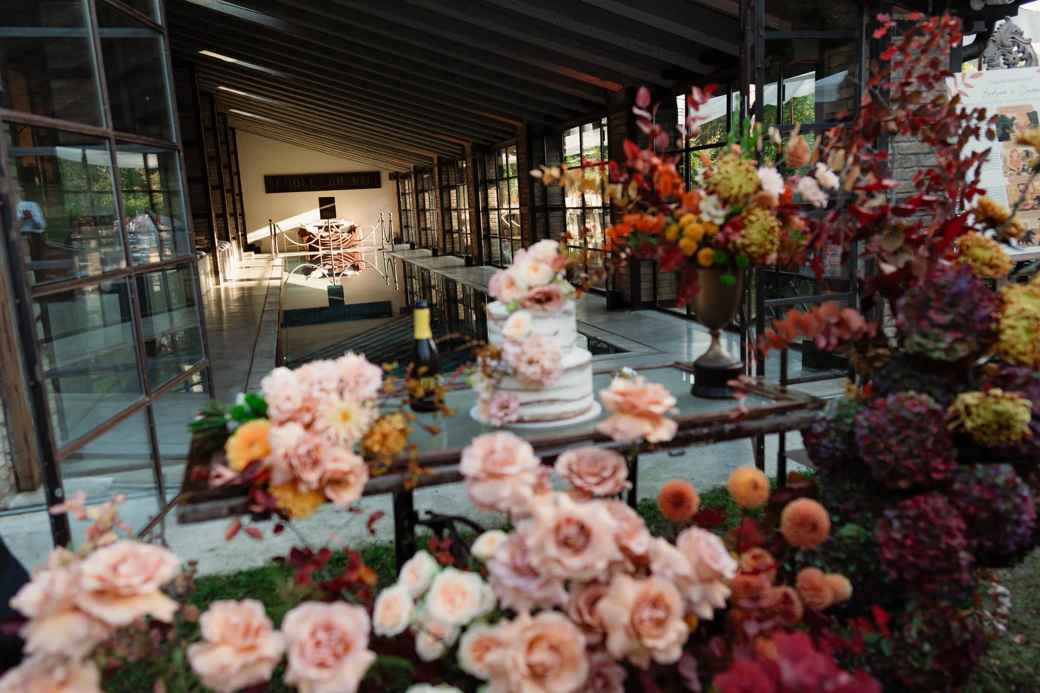 Torta di matrimonio decorata, accompagnata da una confezione di champagne con dettaglio di fiori colorati e vasi con composizioni floreali color pesca, arancio e rosa. Ambiente interno con elementi di arredamento moderno.