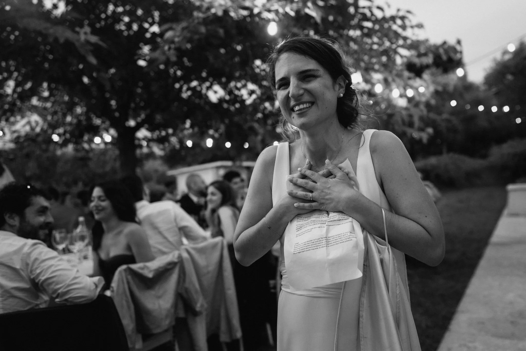 Una donna sorridente con le mani sul cuore durante una cerimonia di matrimonio all'aperto in serata, alberi e luci decorative.