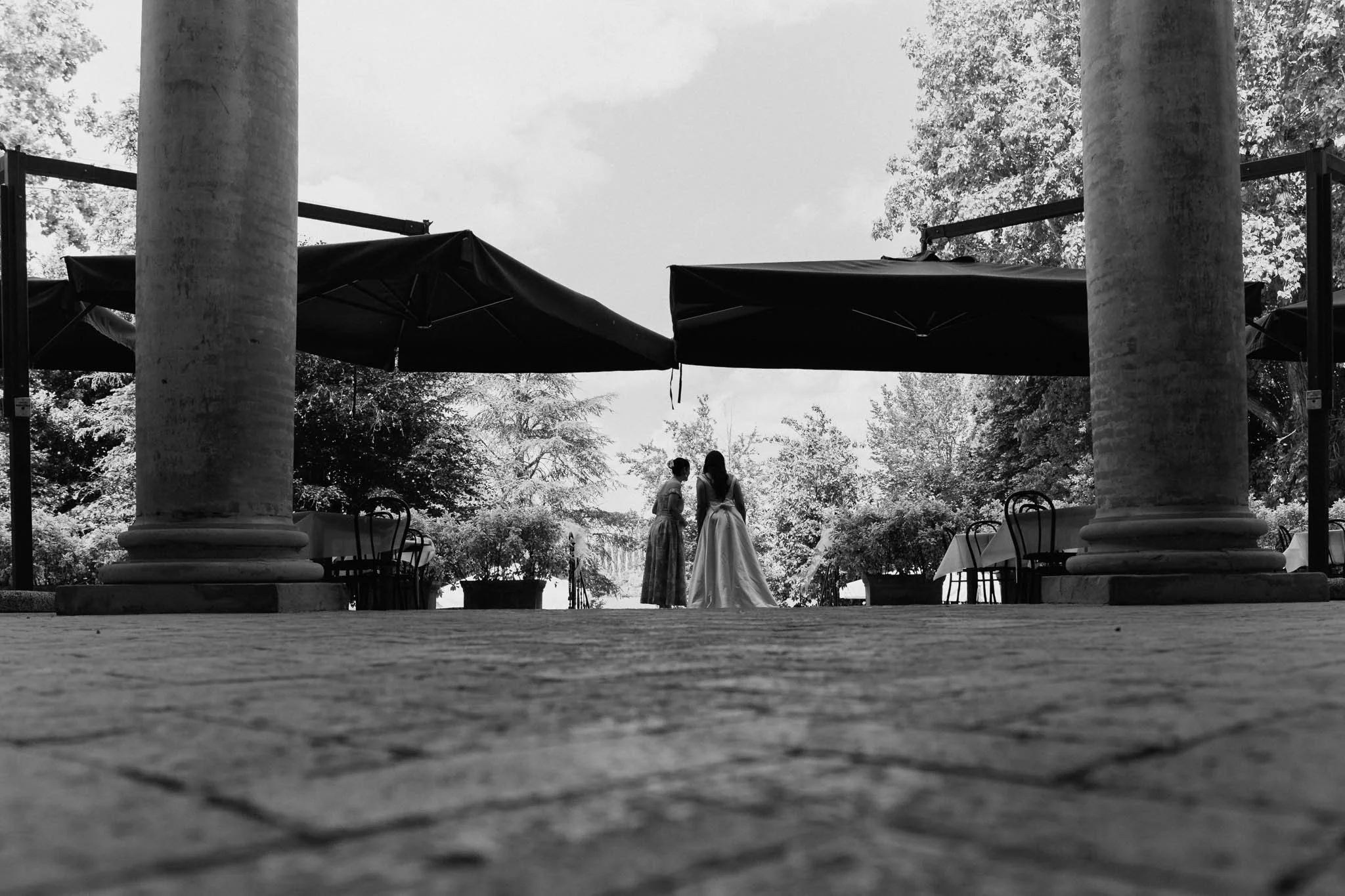 Due donne, una in abito da sposa e una in abito lungo, si trovano sotto un grande ombrello nero su una terrazza con colonne, circondate da alberi e mobili da giardino.