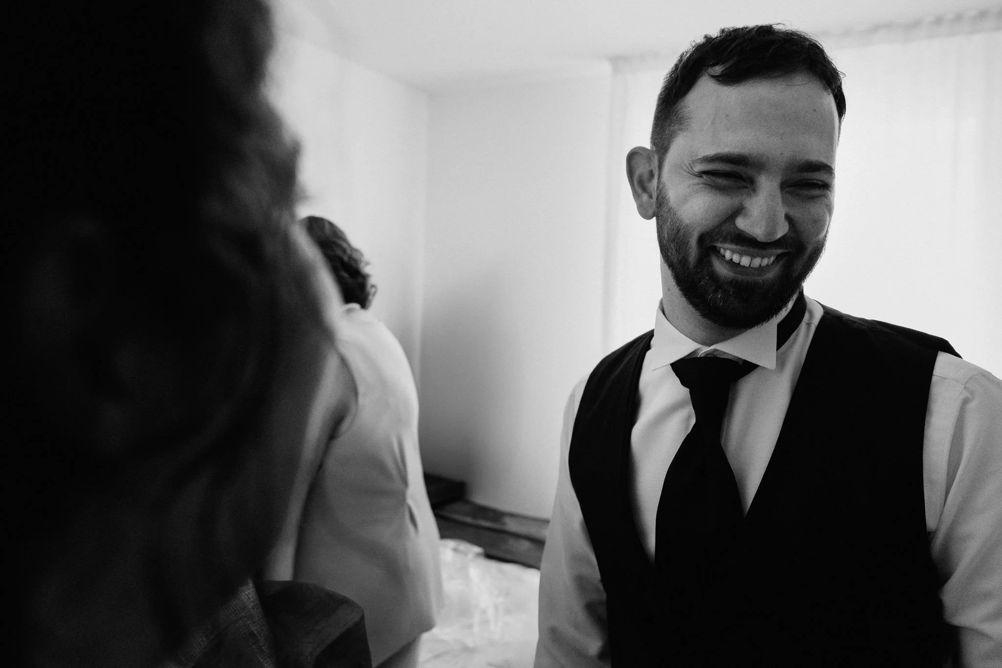 Uomo sorridente in abito elegante con camicia bianca, giacca nera e cravatta, in un ambiente interno, in bianco e nero.