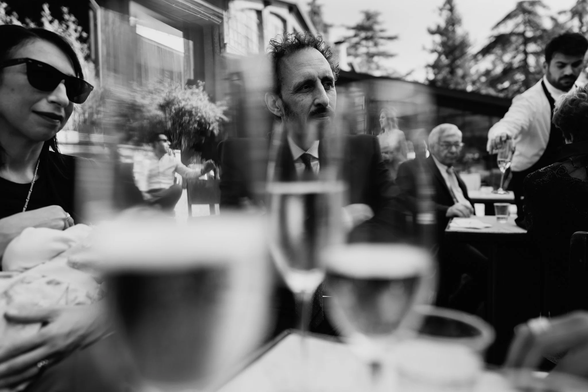 Immagine in bianco e nero di un gruppo di persone sedute a un tavolo durante un evento sociale, con alcuni bicchieri di champagne in primo piano e uno sfondo di alberi e edifici.