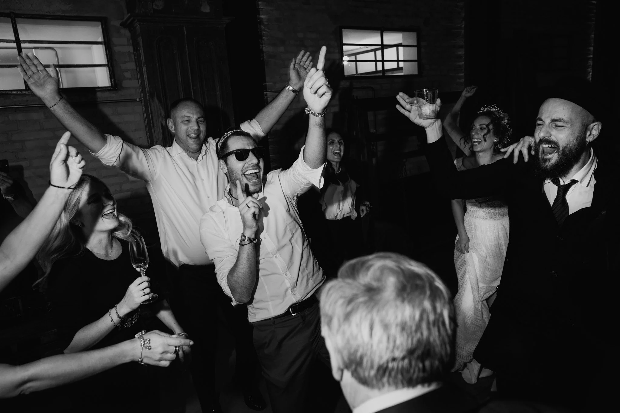 Gente che si diverte e balla a una festa, alcuni indossano vestiti eleganti, alcuni con occhiali da sole e uno con un cappello, in un'atmosfera di festa e allegria.