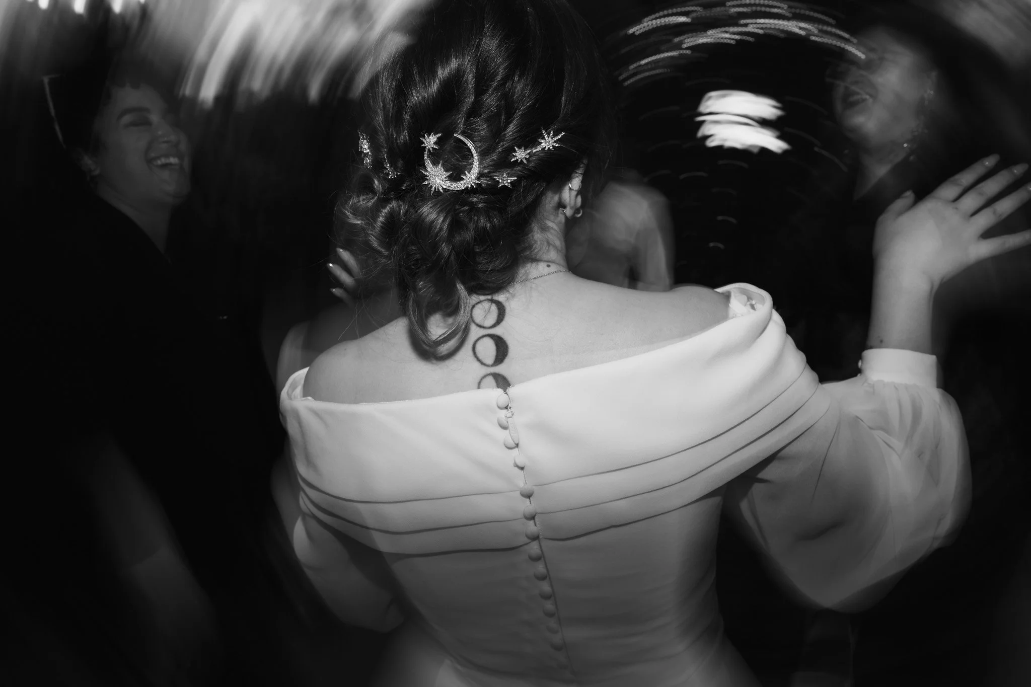 Donna con capelli ricci, decorazioni a forma di luna e stelle, indossa un abito bianco e si trova al centro di una festa o ballo, circondata da altre persone che ridono e si divertono.