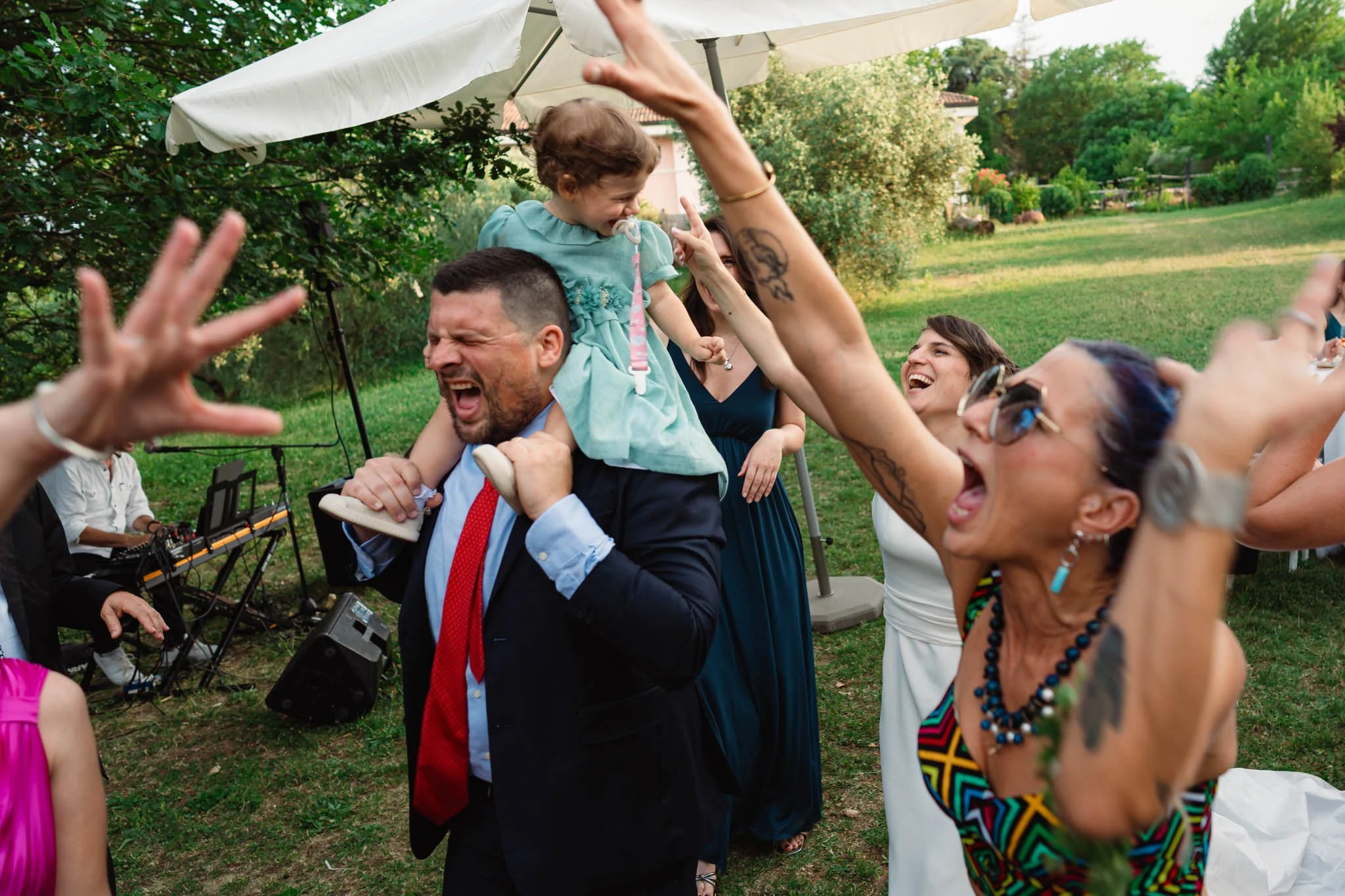 Gruppo di persone che si divertono a un evento all'aperto, con musica e festeggiamenti a un matrimonio, sotto un ombrellone in un prato verde.
