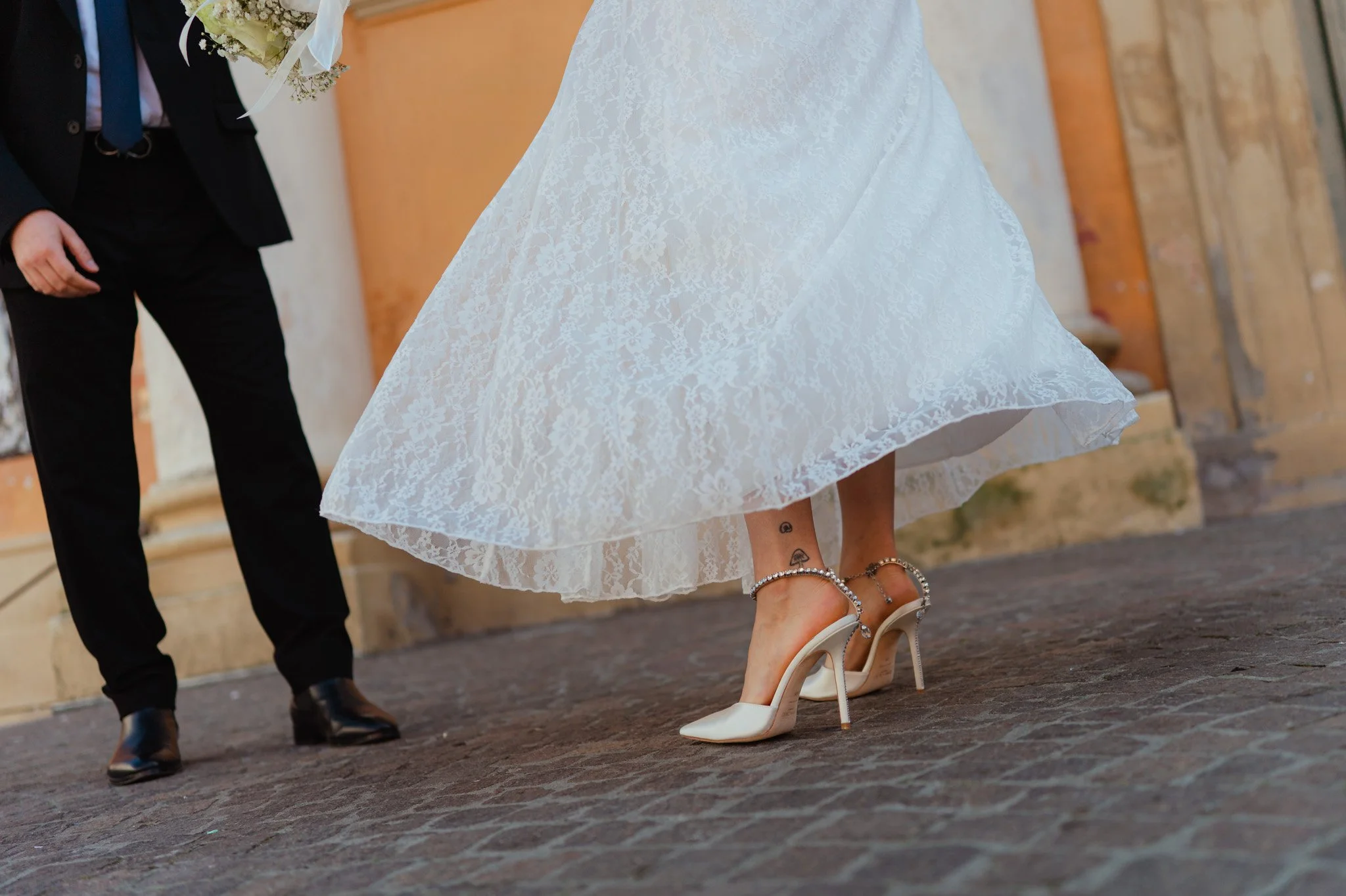 Dettaglio di una donna che indossa scarpe da tacco alte e un abito bianco di pizzo, mentre un uomo in abito nero la guarda.