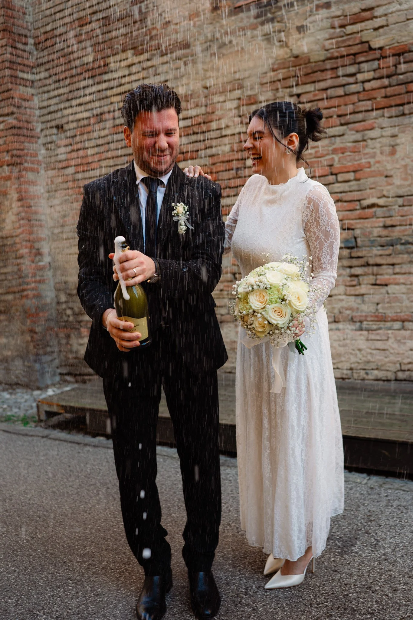 Nozze con champagne sotto la pioggia, sposo e sposa sorridenti, sposo con giacca nera, sposa con abito in pizzo e bouquet di fiori bianchi, muro di mattoni sullo sfondo.