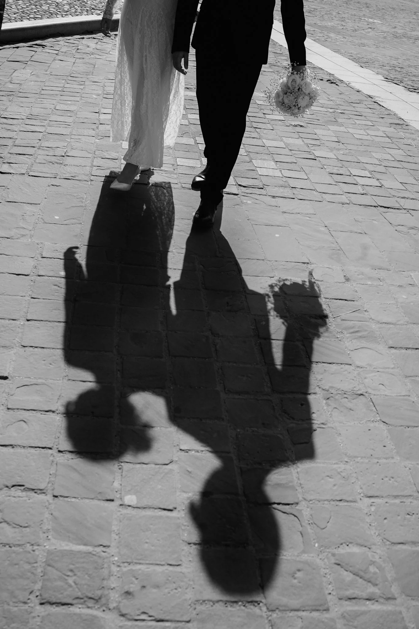 Un uomo e una donna che camminano su un marciapiede di pietra, con le ombre proiettate sul pavimento, la donna tiene un bouquet di fiori.
